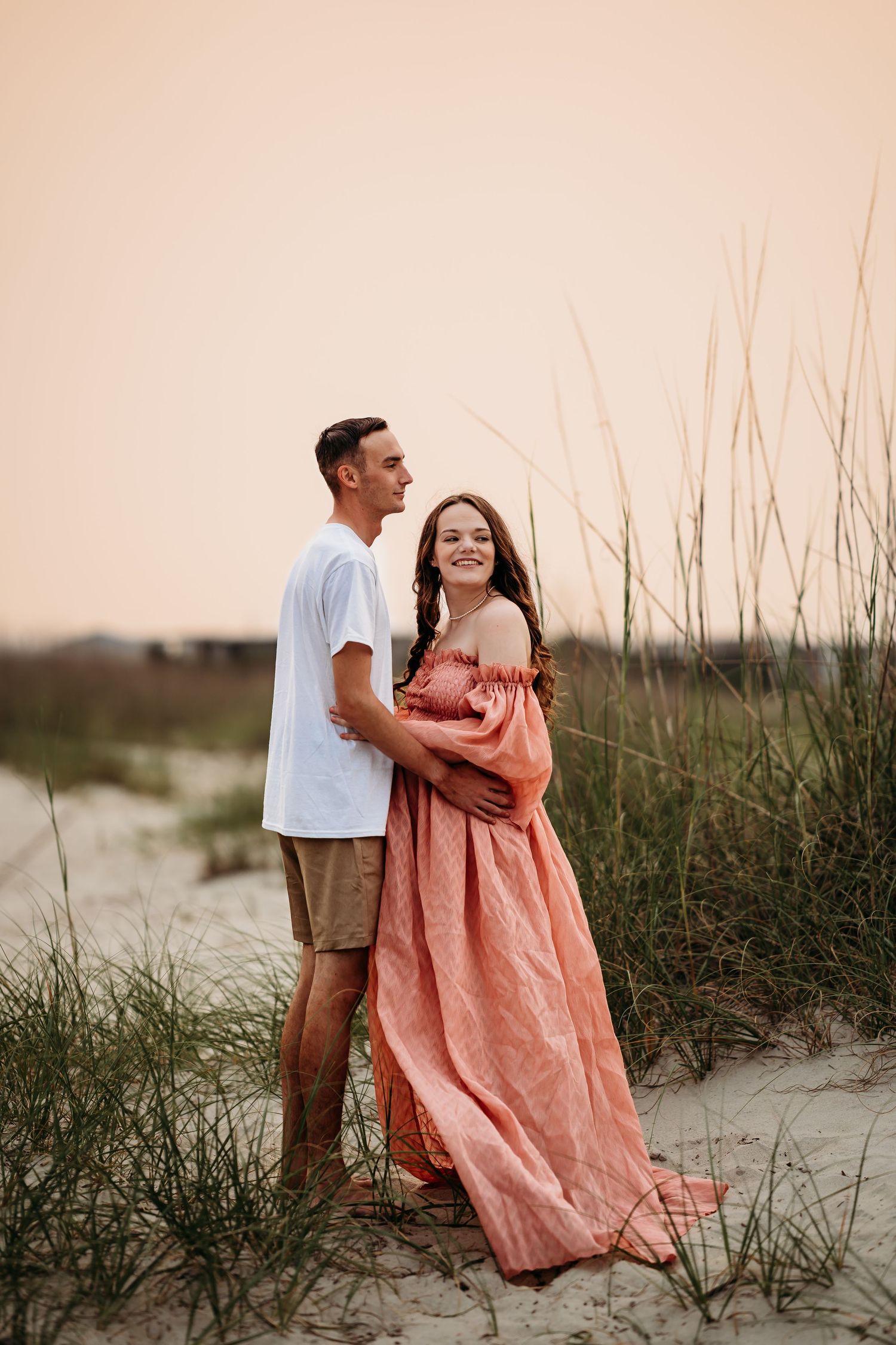 Couple embracing on beach dunes at sunset, pregnant woman in coral gown with partner in white tee.