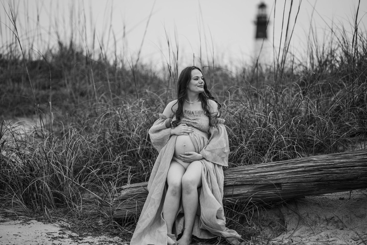 Black and white photo of pregnant woman sitting on driftwood near lighthouse during maternity shoot.