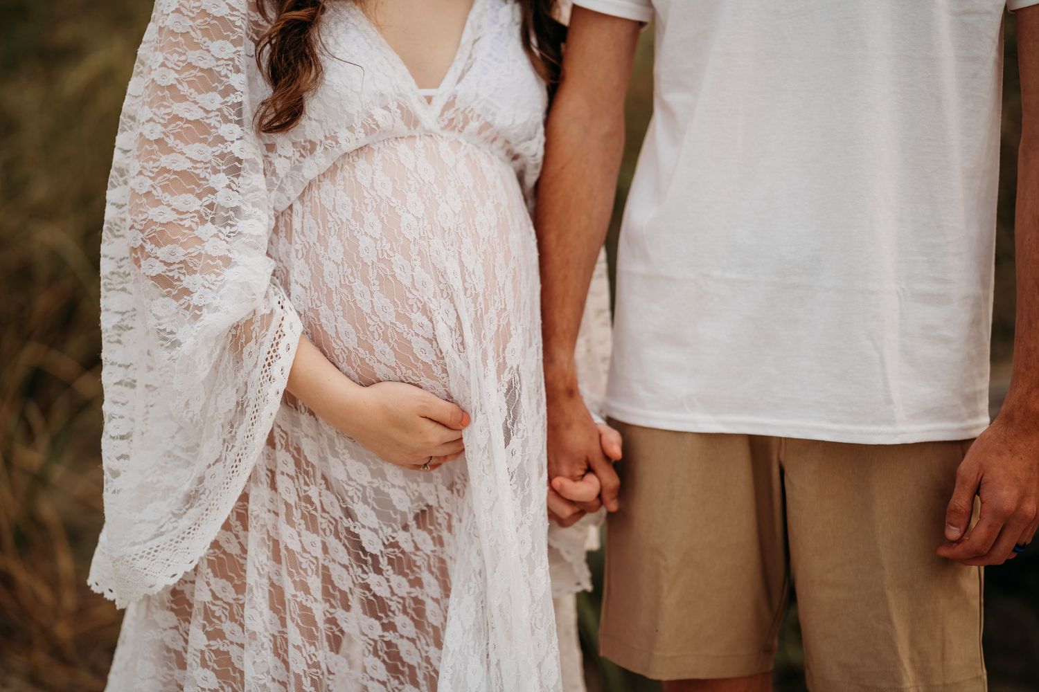 Close-up of pregnant woman in white lace dress holding hands with partner on beach.
