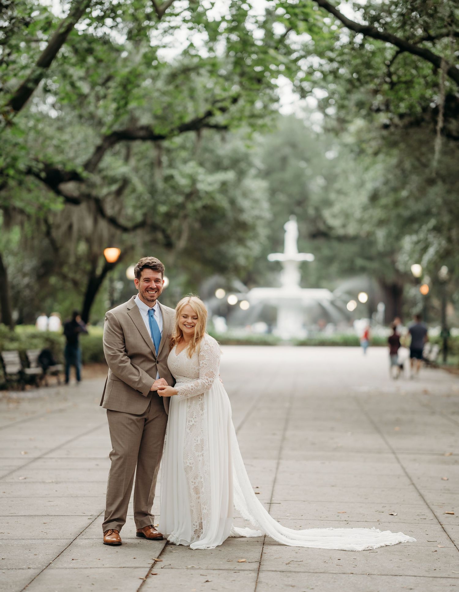 Smiling bride and groom hold hands on Forsyth Park walkway with iconic fountain in background.
