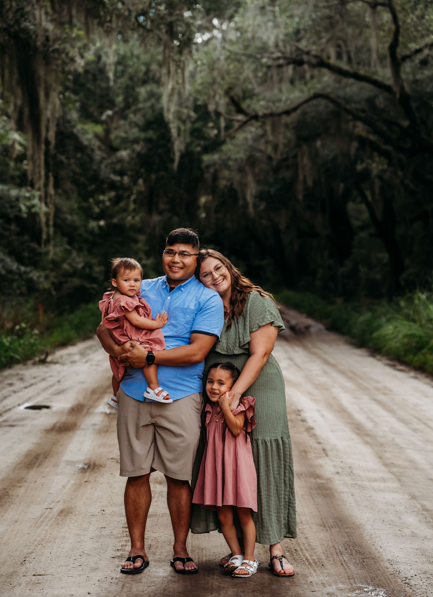 Happy family of four posing on moss-lined dirt road surrounded by lush green trees.