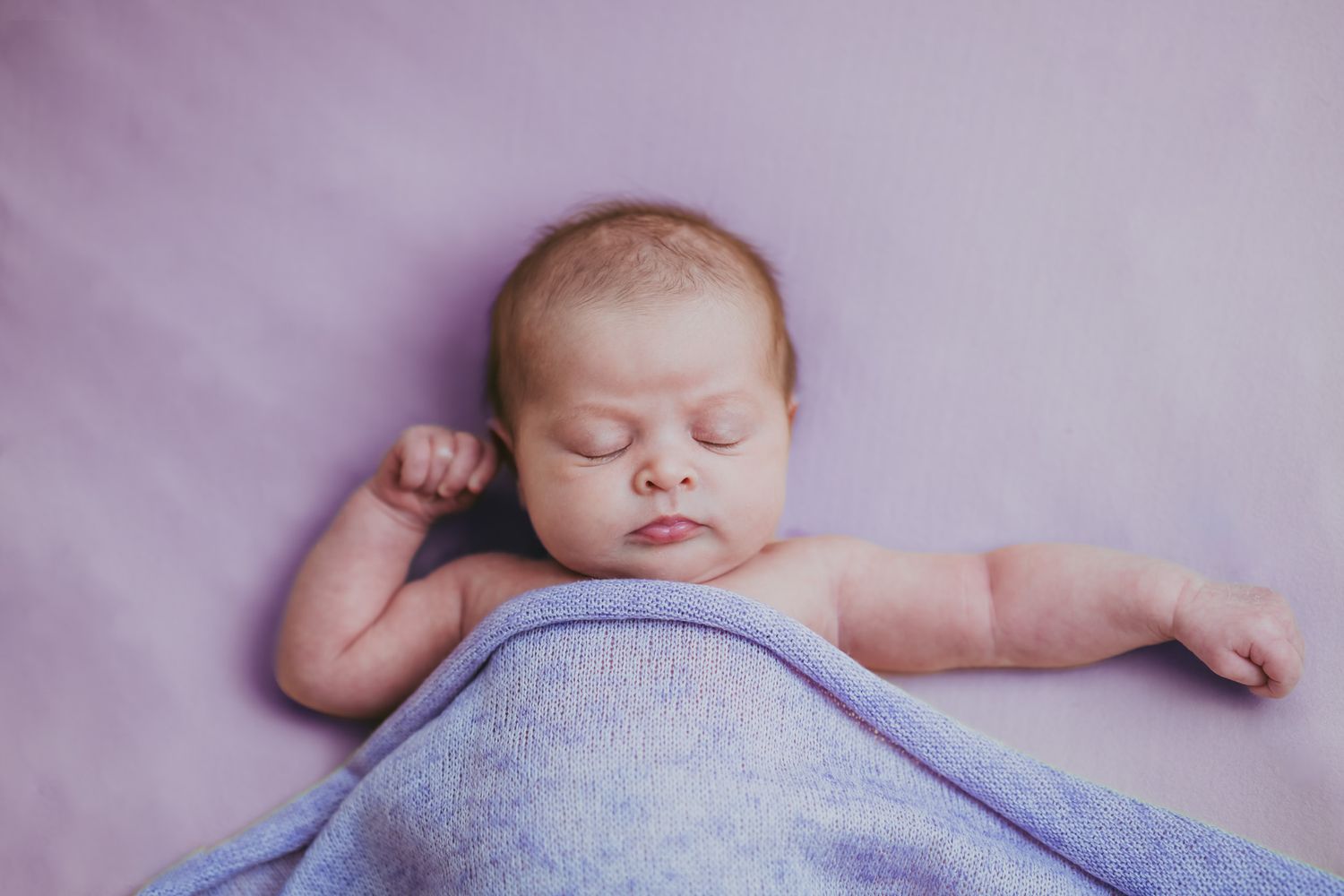 Peaceful newborn baby sleeping on purple blanket with arms raised in serene portrait photo.