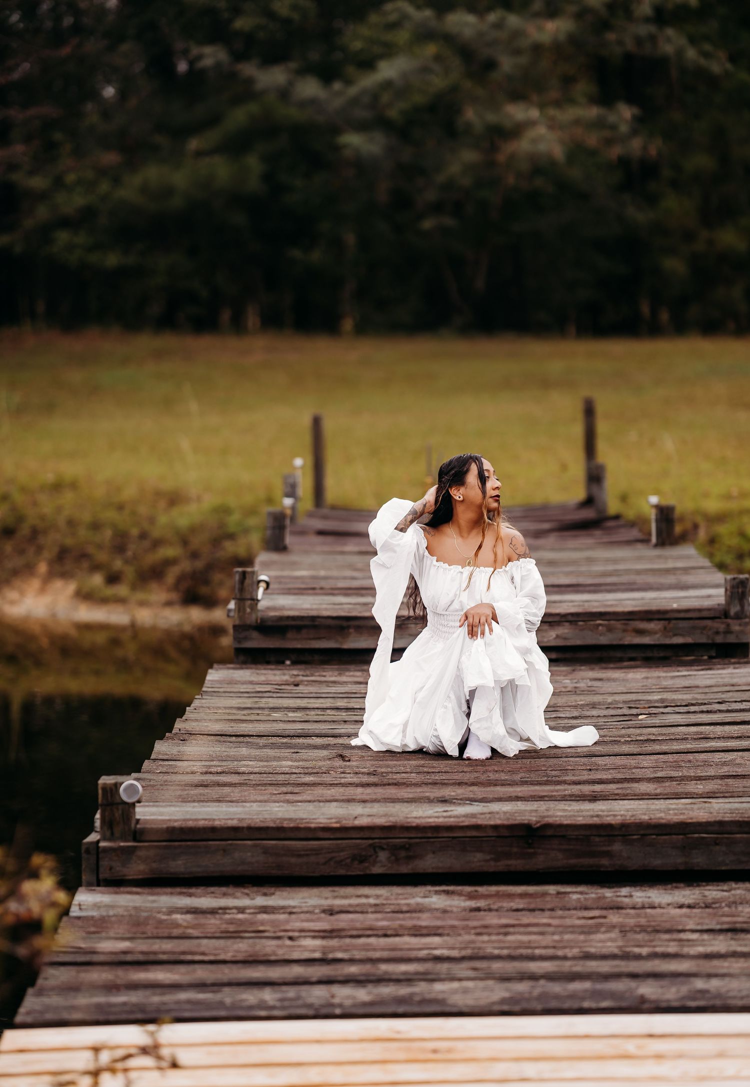 Joyful bride in white dress sits laughing on a wooden dock beside a peaceful pond.