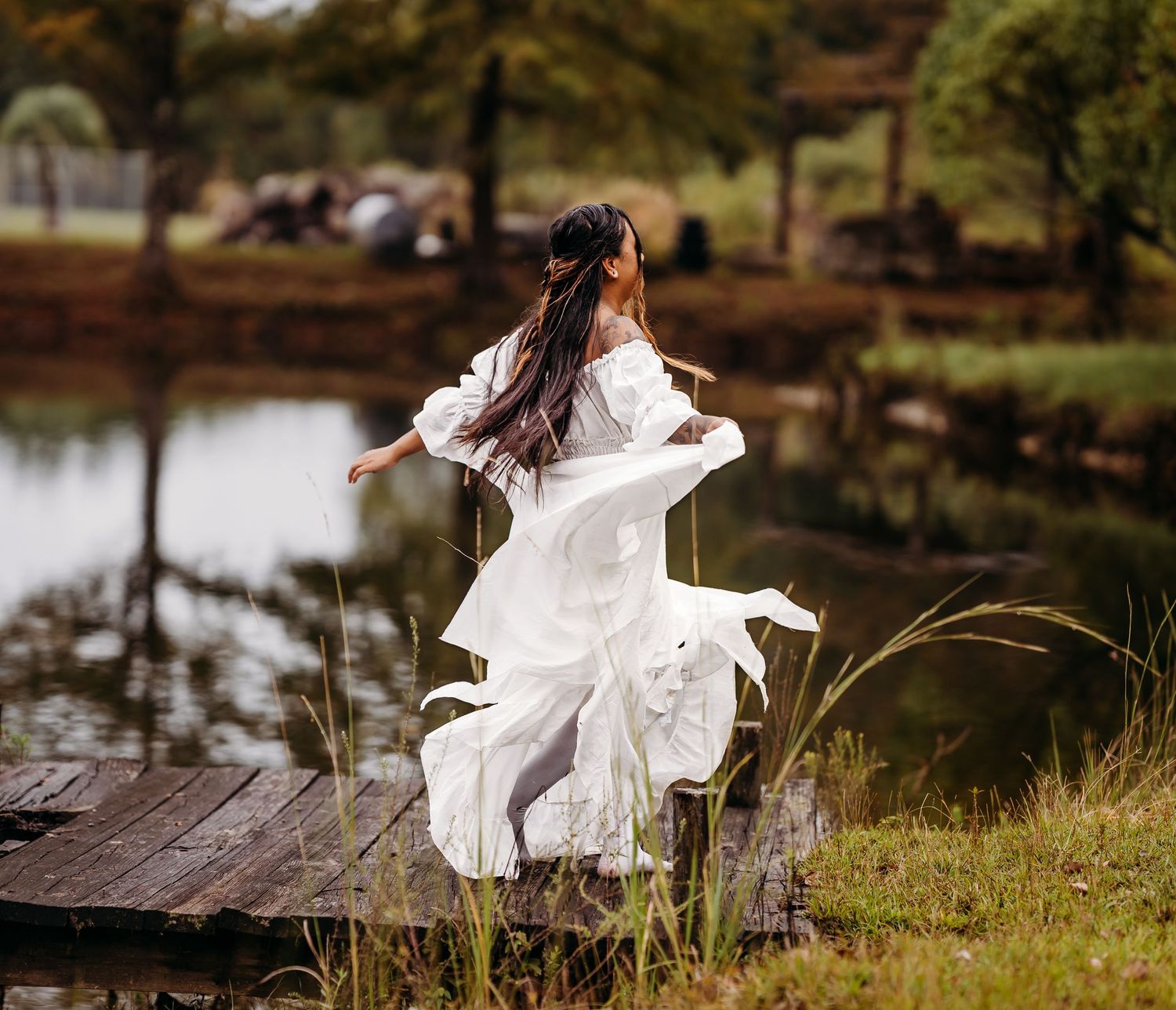Woman in flowing white dress dances freely on wooden pier surrounded by lush greenery and pond.