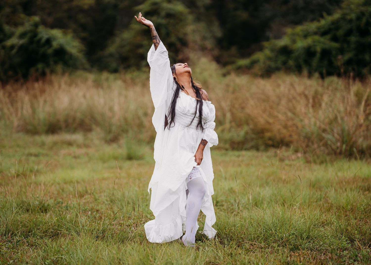 Woman in white dress performing expressive pose with arm raised in open grassy meadow.