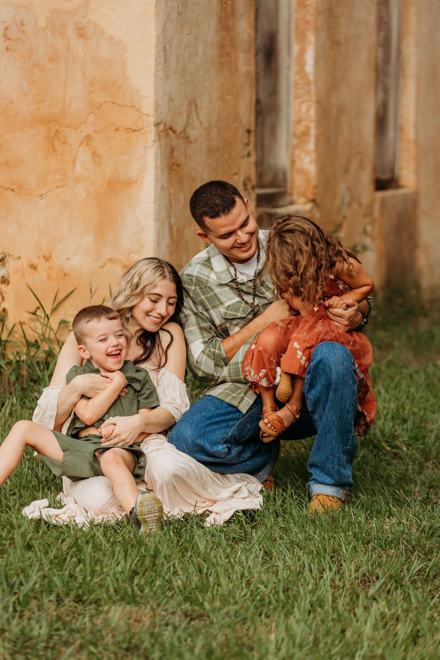Happy family of four sitting on grass, laughing together outdoors near a rustic wall.