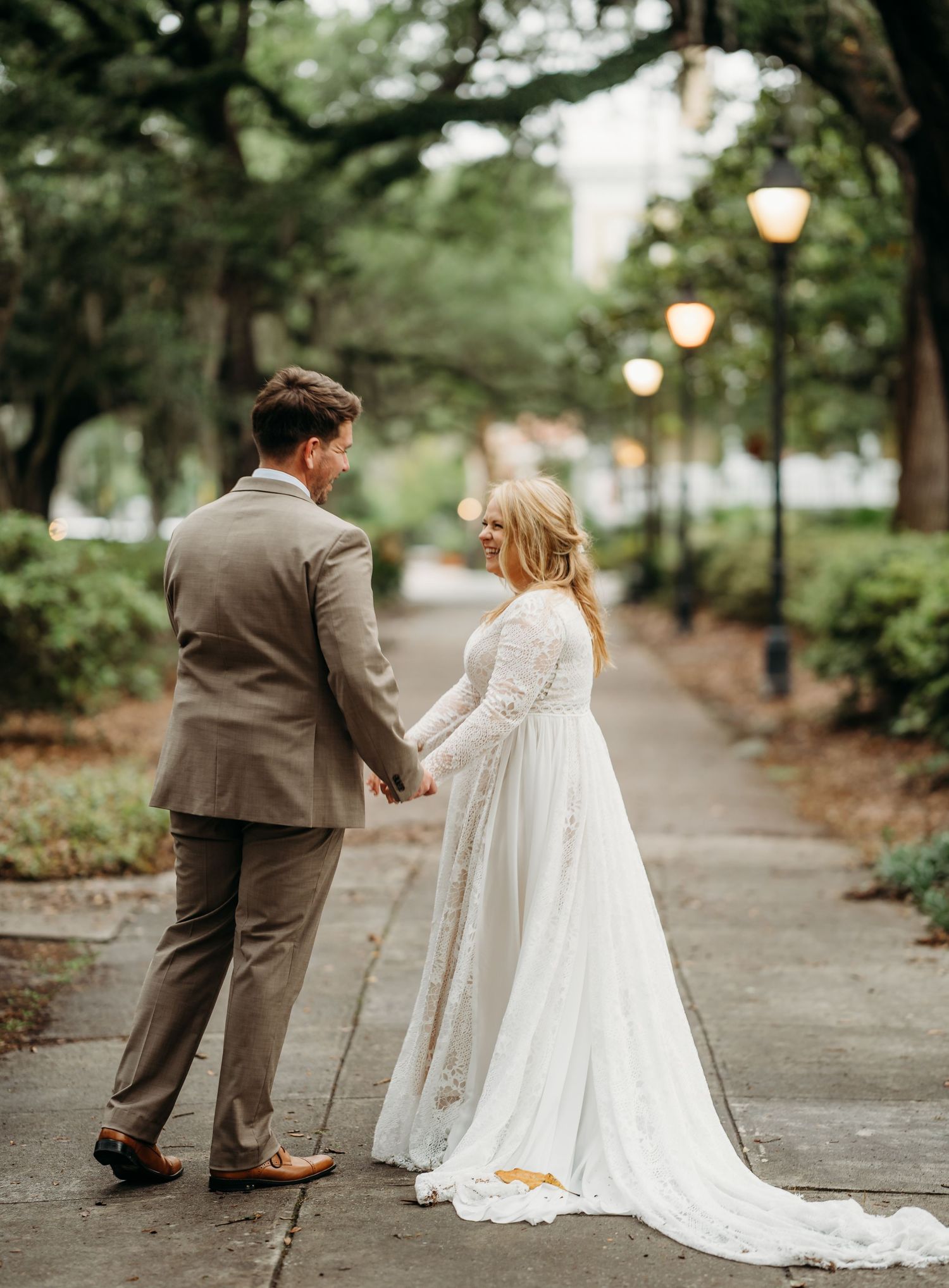 Bride in flowing white lace gown and groom in tan suit walk hand-in-hand down a scenic tree-lined sidewalk at dusk.