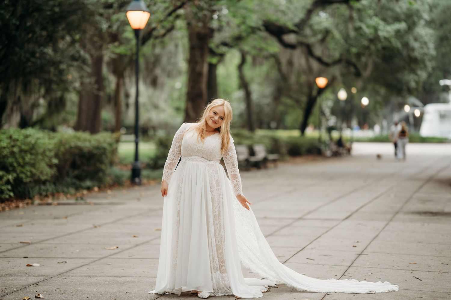 Bride in lace long-sleeve wedding gown poses gracefully on tree-lined park pathway.