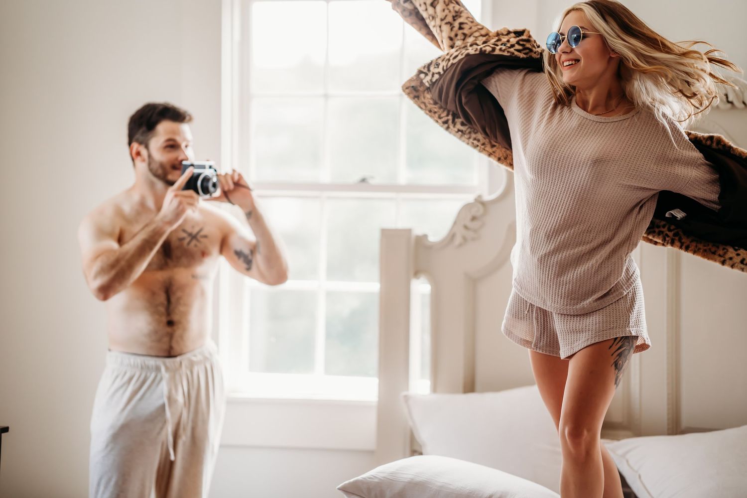 Man photographing playful woman swinging leopard coat on a bright bedroom bed.