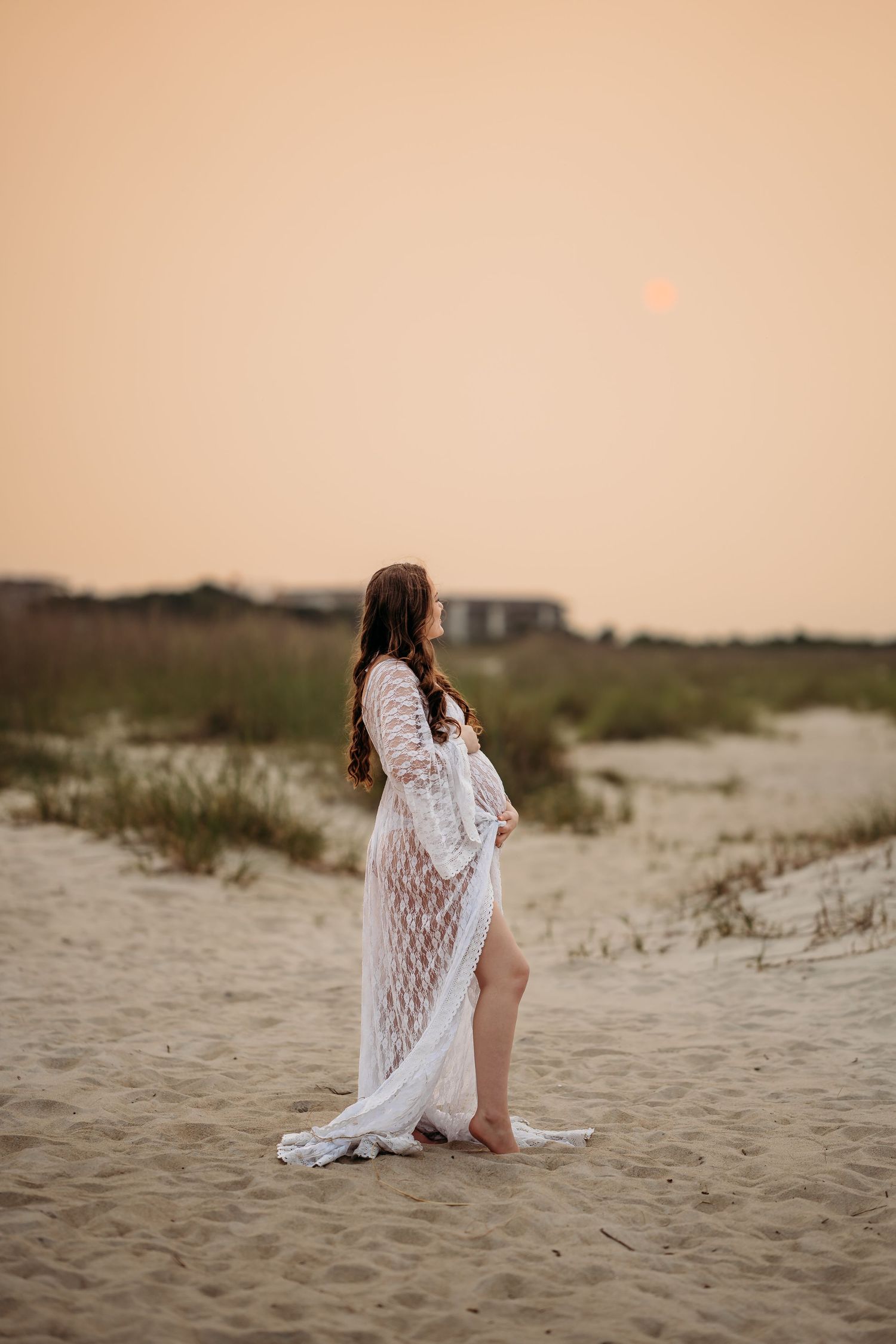 Pregnant woman in white lace maternity gown standing on sandy beach at golden sunset.
