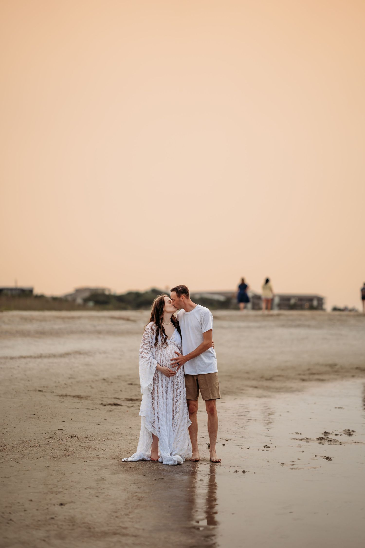 Couple sharing a tender moment kissing on sandy beach shoreline during warm golden sunset.