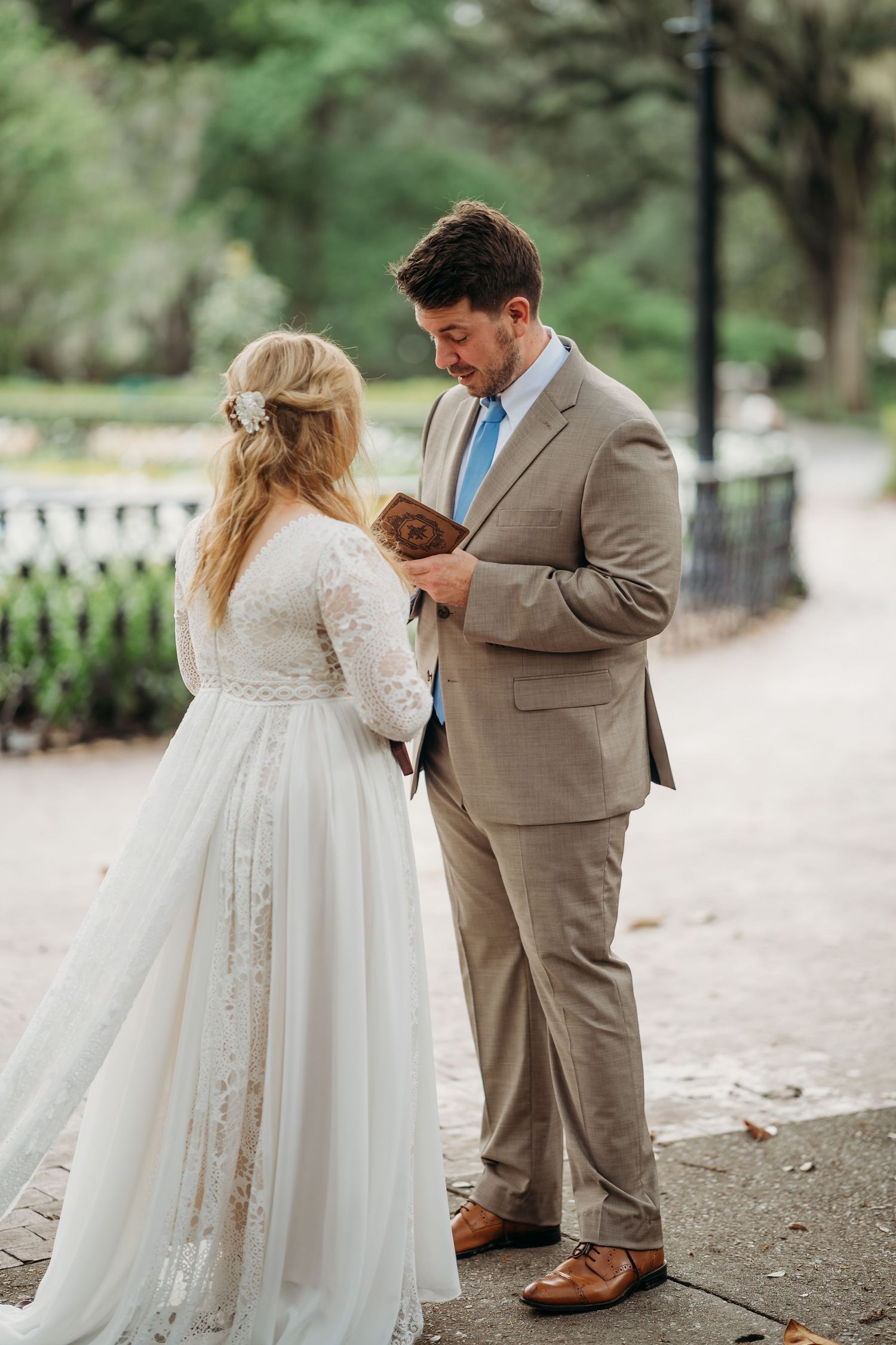 Groom in tan suit reads vows from small book while standing with bride in lace gown outdoors.