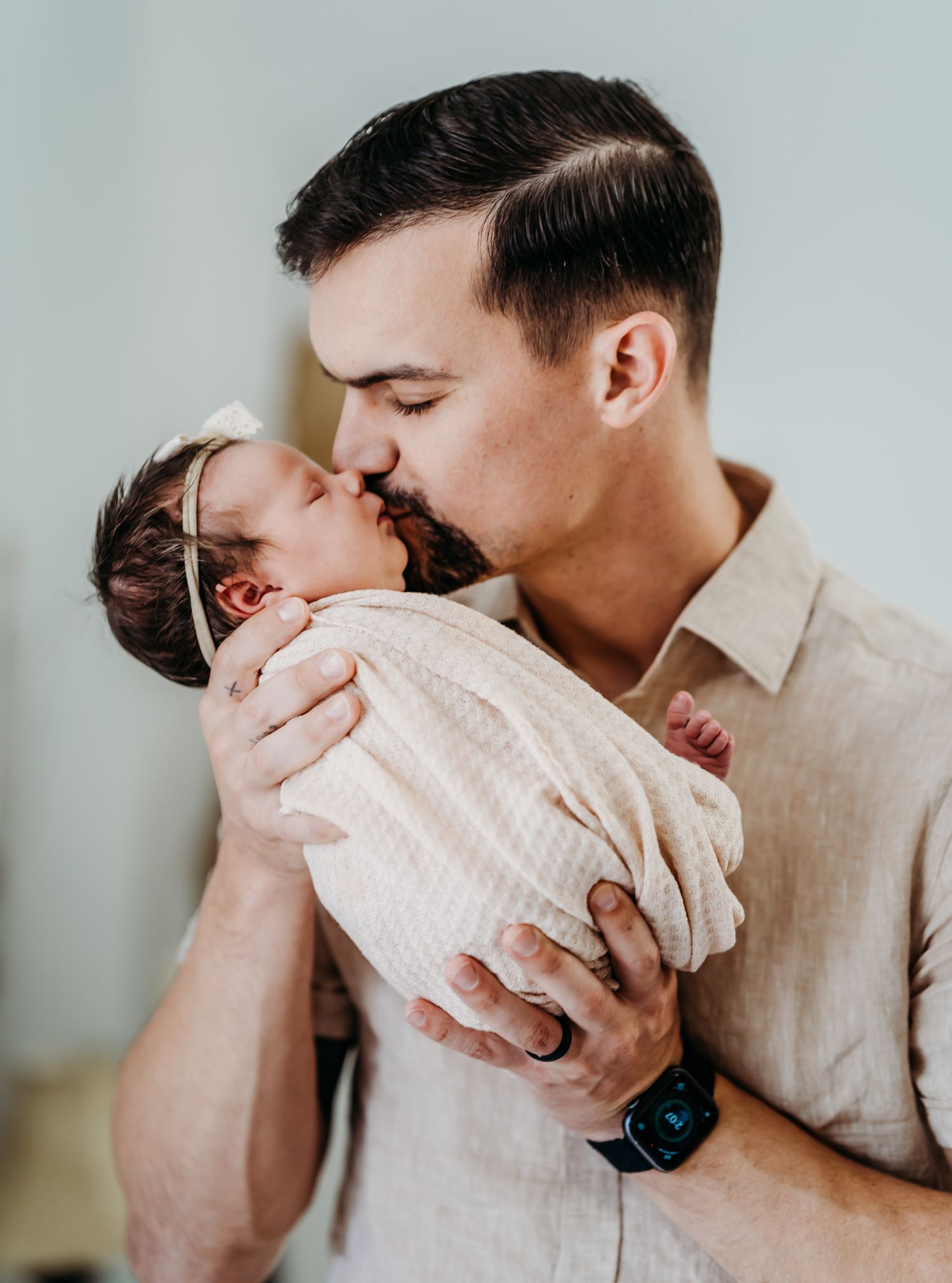 Loving father kisses his swaddled newborn baby girl gently on the forehead indoors.