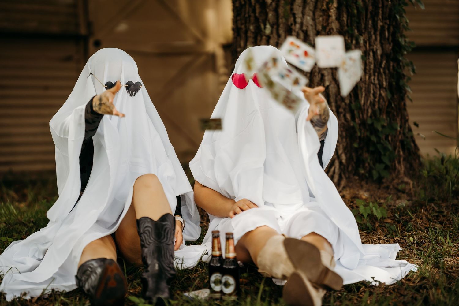 Two people dressed as ghosts in white sheets sitting under a tree with beer bottles.
