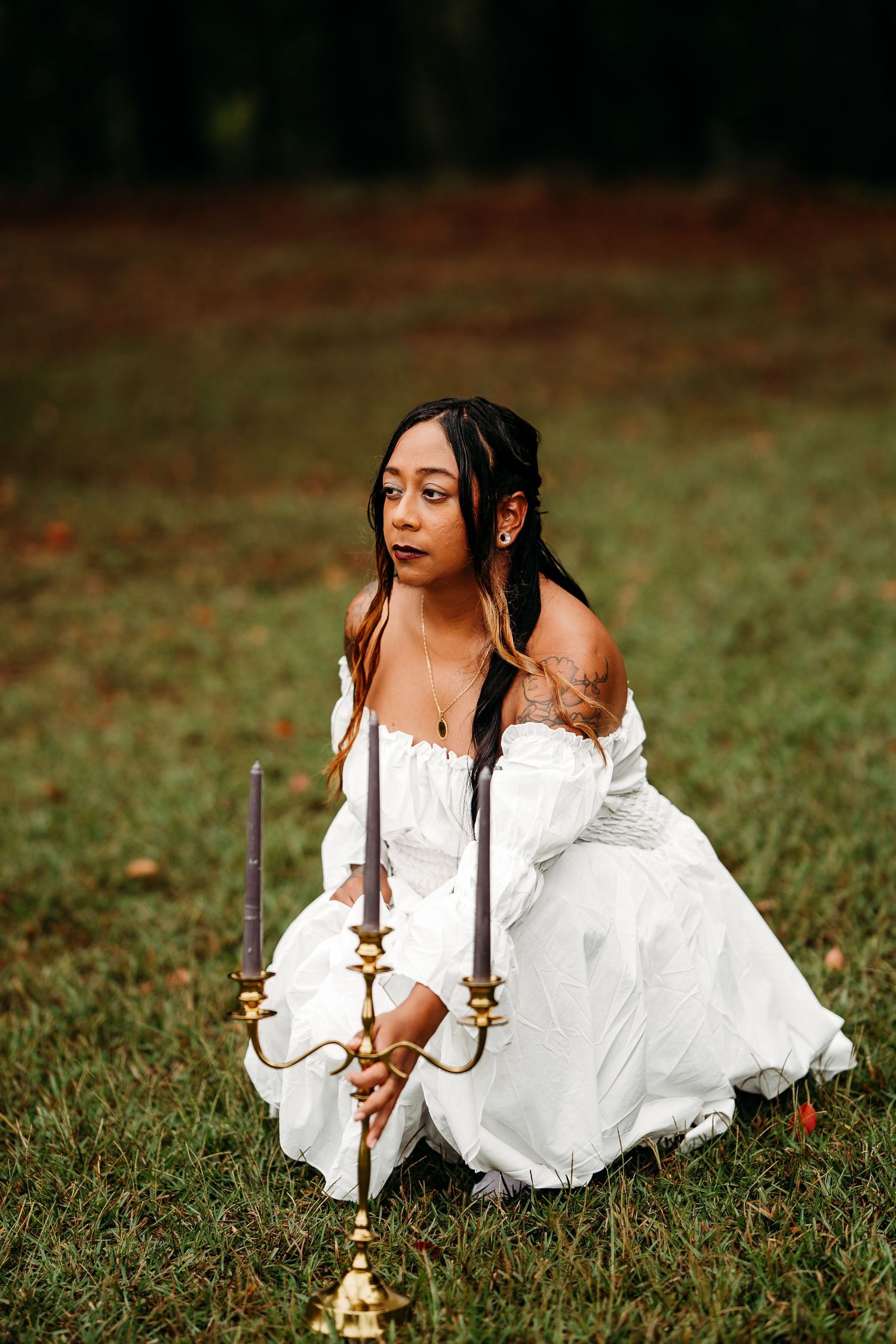 Young woman in white off-shoulder gown kneels on grass beside gold three-arm candelabra in outdoor park.