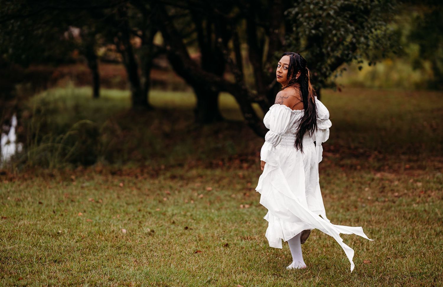 Woman in flowing white dress walking gracefully across green field with autumn trees behind.