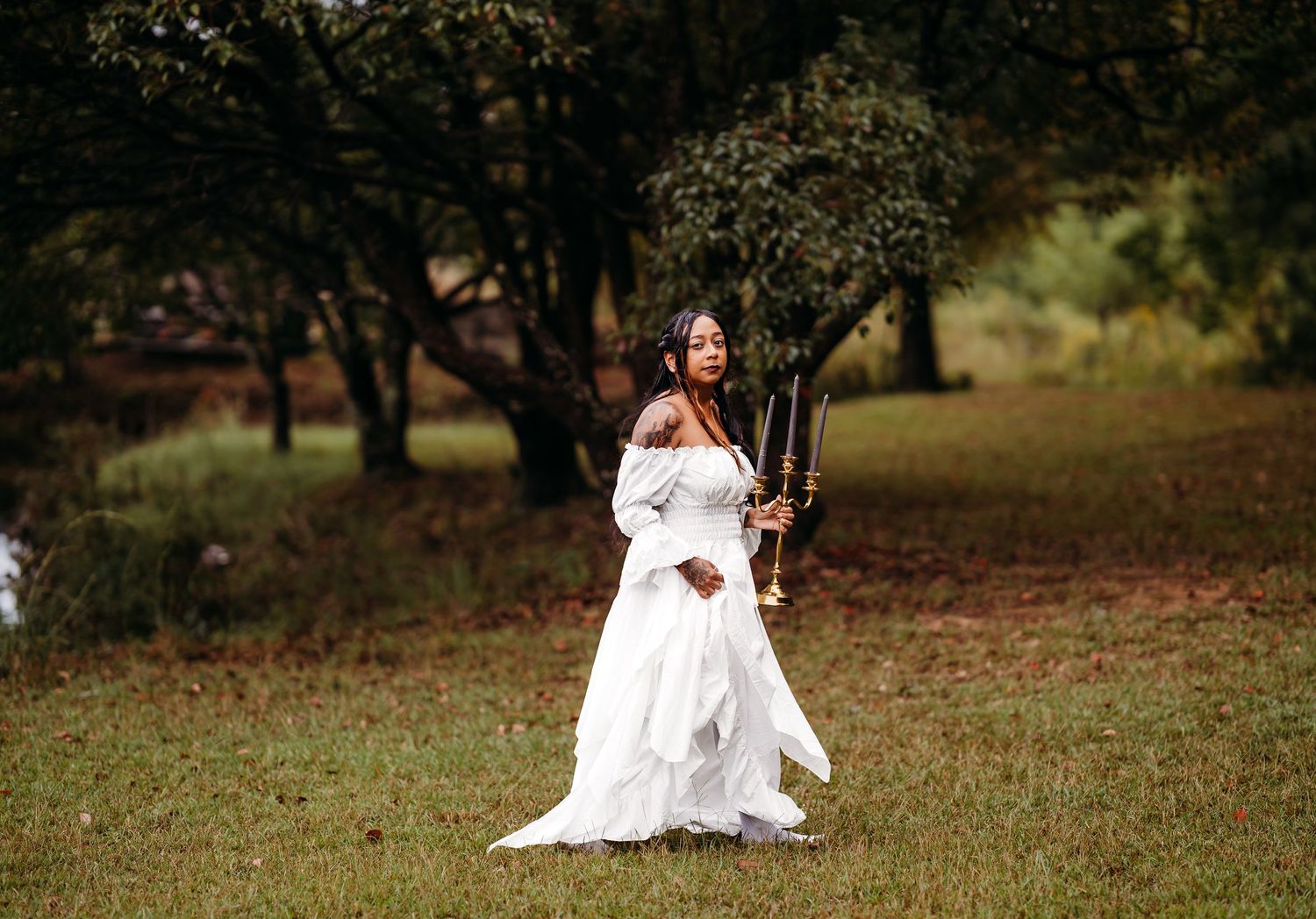 Woman in white off-shoulder dress stands in open grassy field holding gold candelabra among autumn trees.