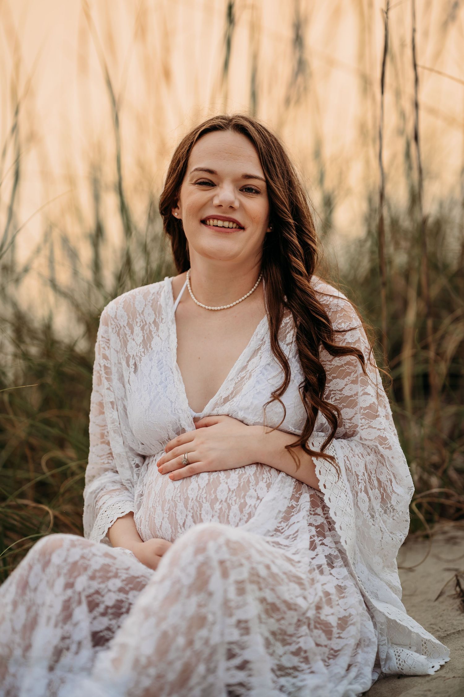 Happy pregnant woman in white lace dress sitting among beach grasses during warm sunset.