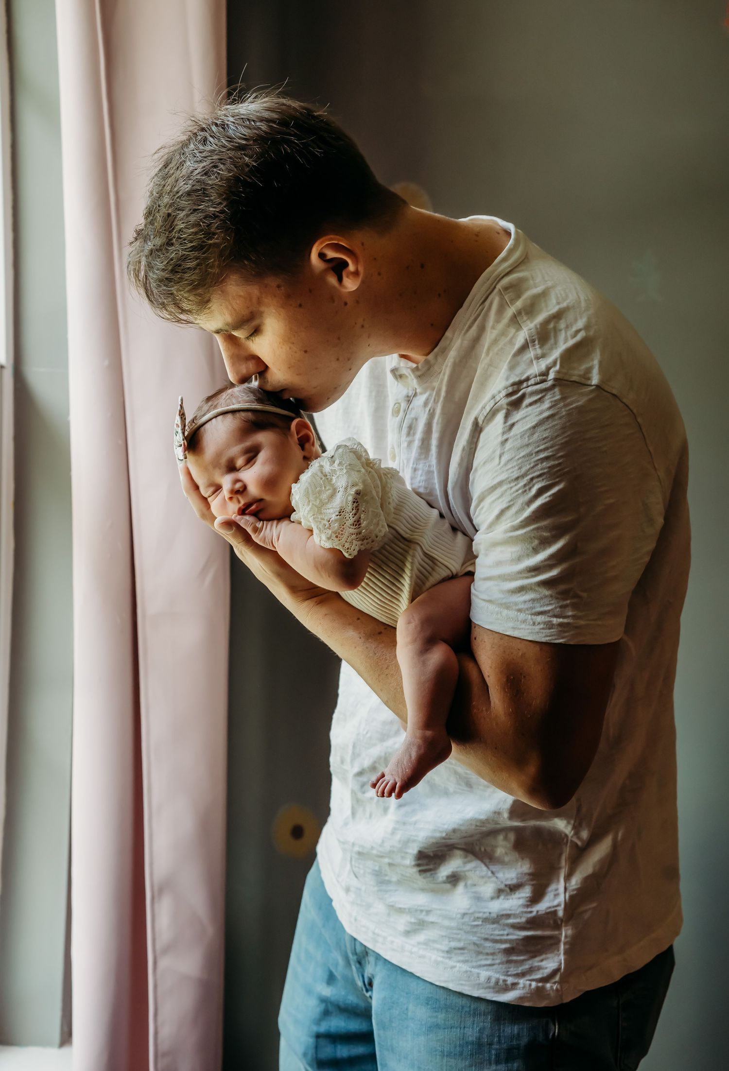 Loving father kissing newborn baby by pink curtained window in soft light.