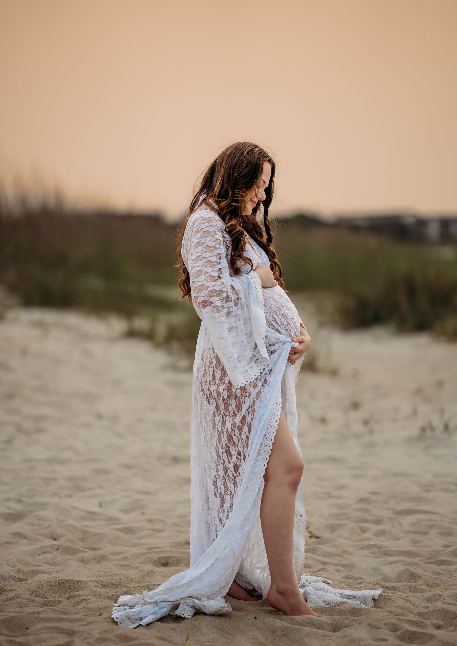 Pregnant woman in flowing white lace gown cradling baby bump on sandy beach at sunset.