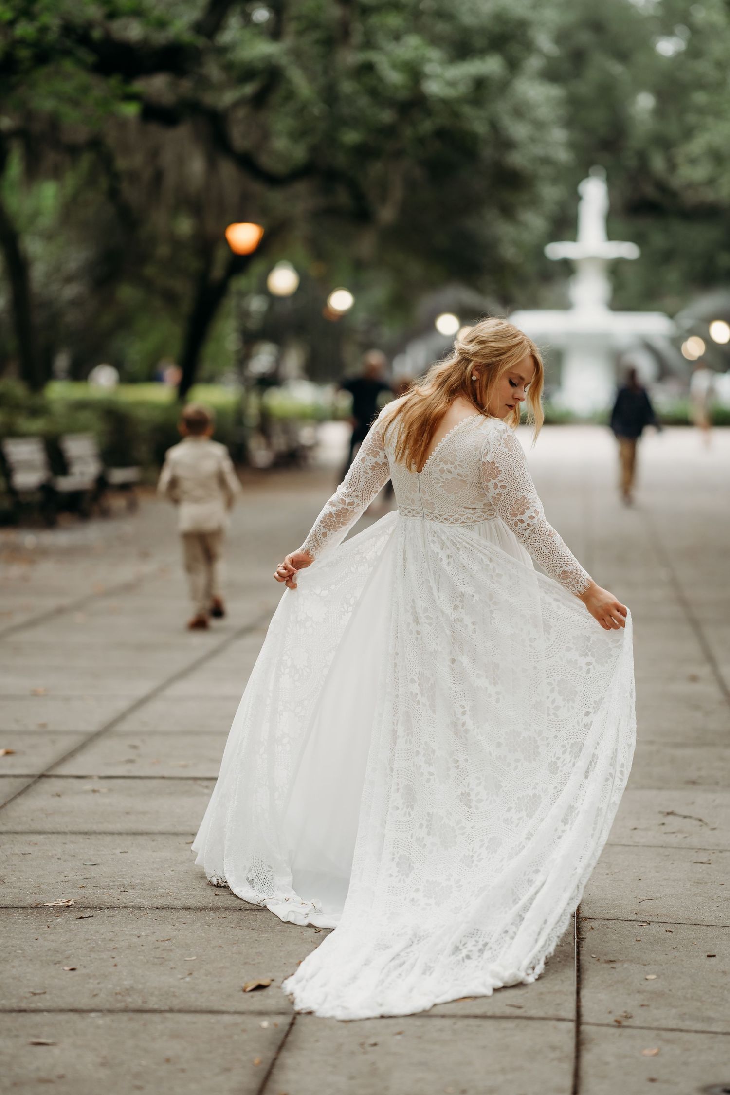 Bride in long-sleeve lace gown playfully swirls skirt on park pathway near fountain.