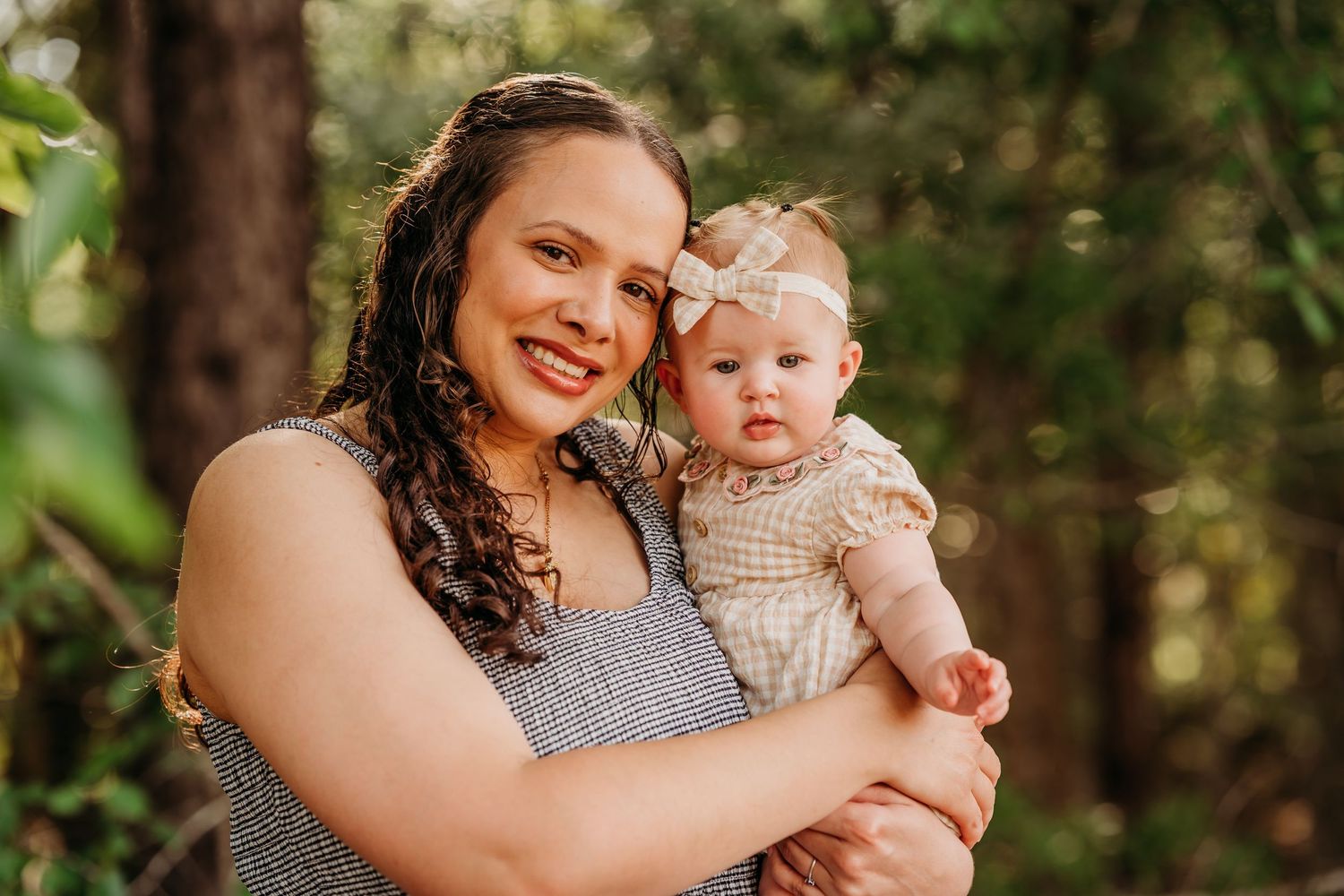 A smiling woman with curly hair holds a cute baby girl wearing a floral headband outdoors among green trees.