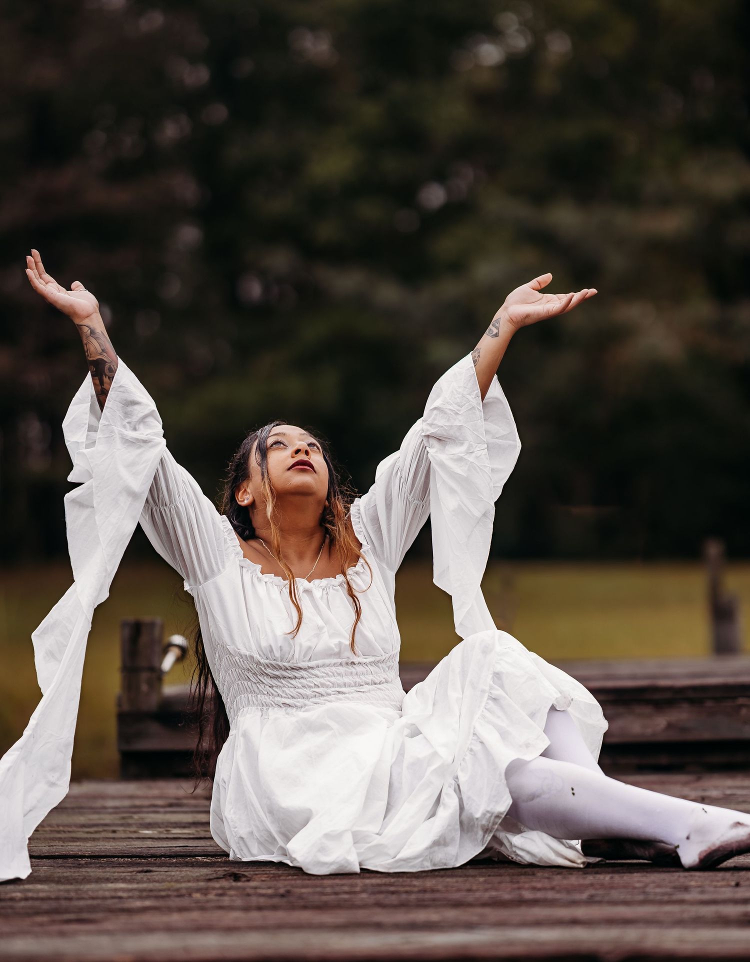 Woman in white dress sitting on wooden dock with arms raised gracefully toward sky.