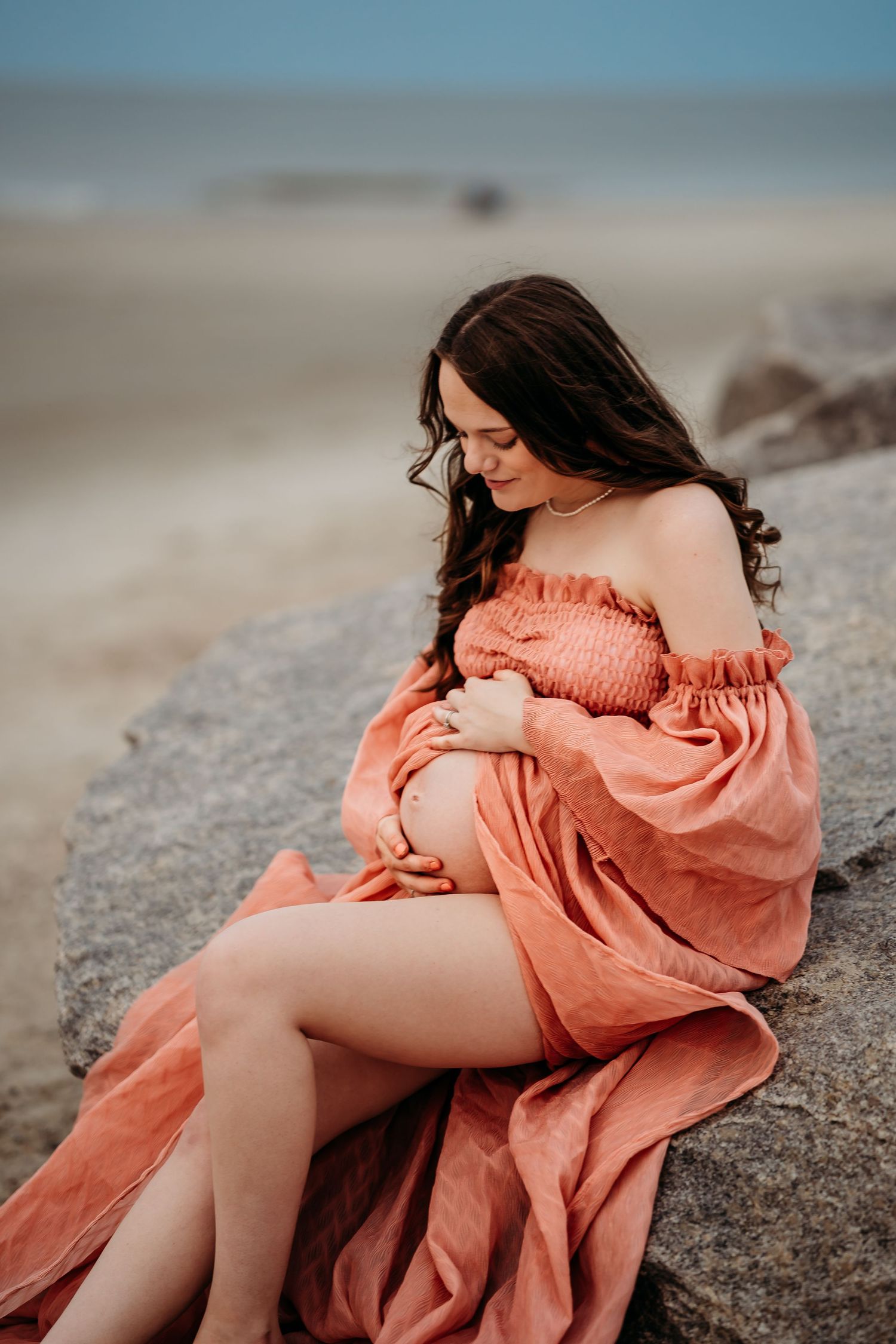 Pregnant woman in coral off-shoulder dress sitting on beach rock, gazing down at her bump.