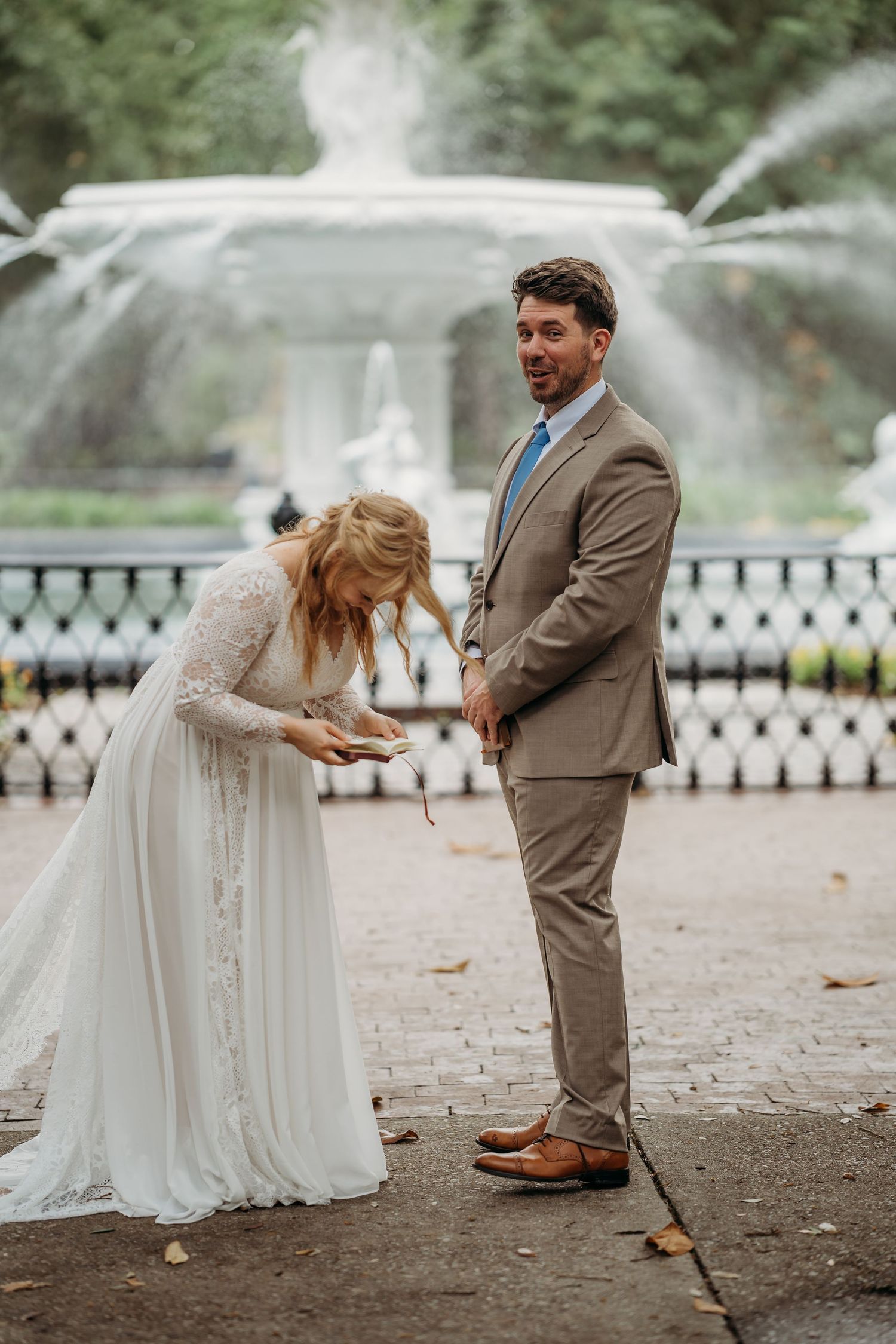 Bride in white lace dress reads vows while groom in tan suit smiles warmly by a grand fountain.