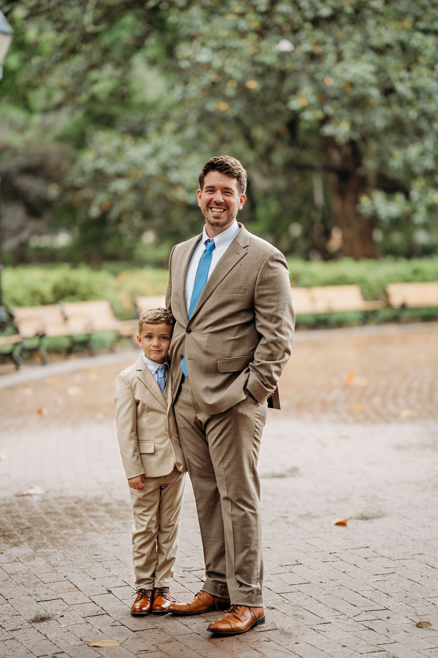 Groom and young ring bearer in matching tan suits posing in outdoor park.