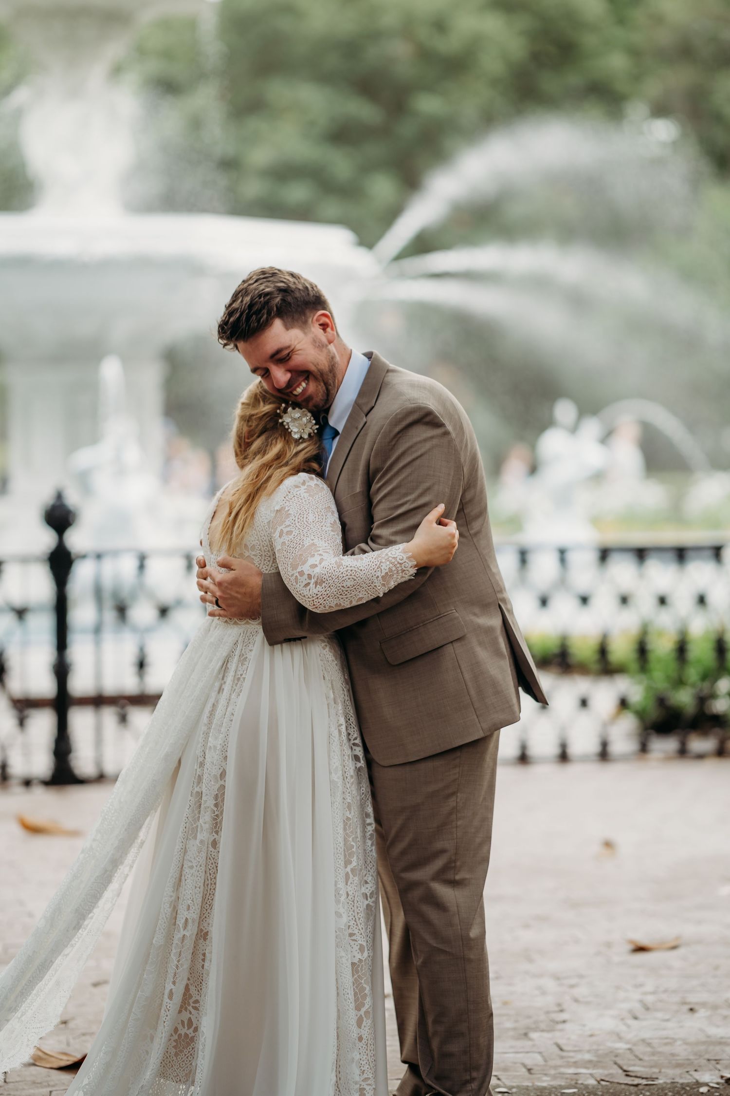 Newlyweds share a warm embrace in front of a grand fountain, both smiling with joy on their wedding day.