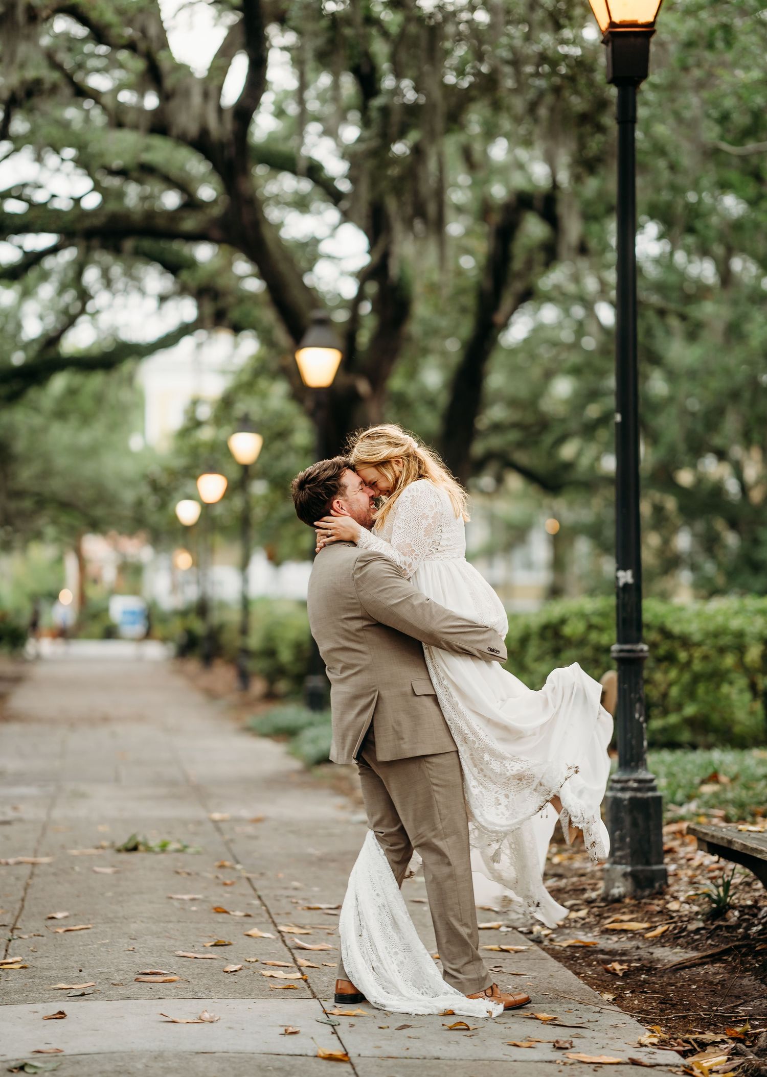 Groom lifts his bride for a kiss beside a glowing lamppost along a scenic tree-lined promenade.