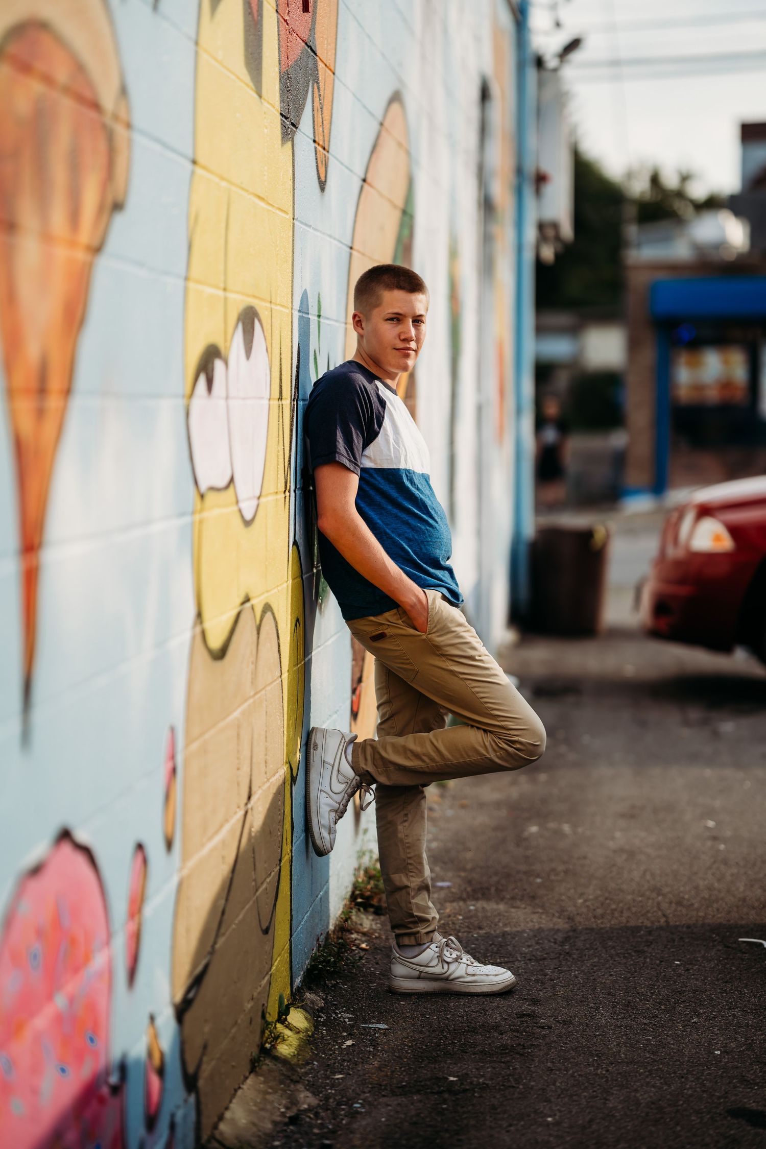 Teen boy in blue shirt and khaki pants leans against colorful graffiti wall in urban alley setting.