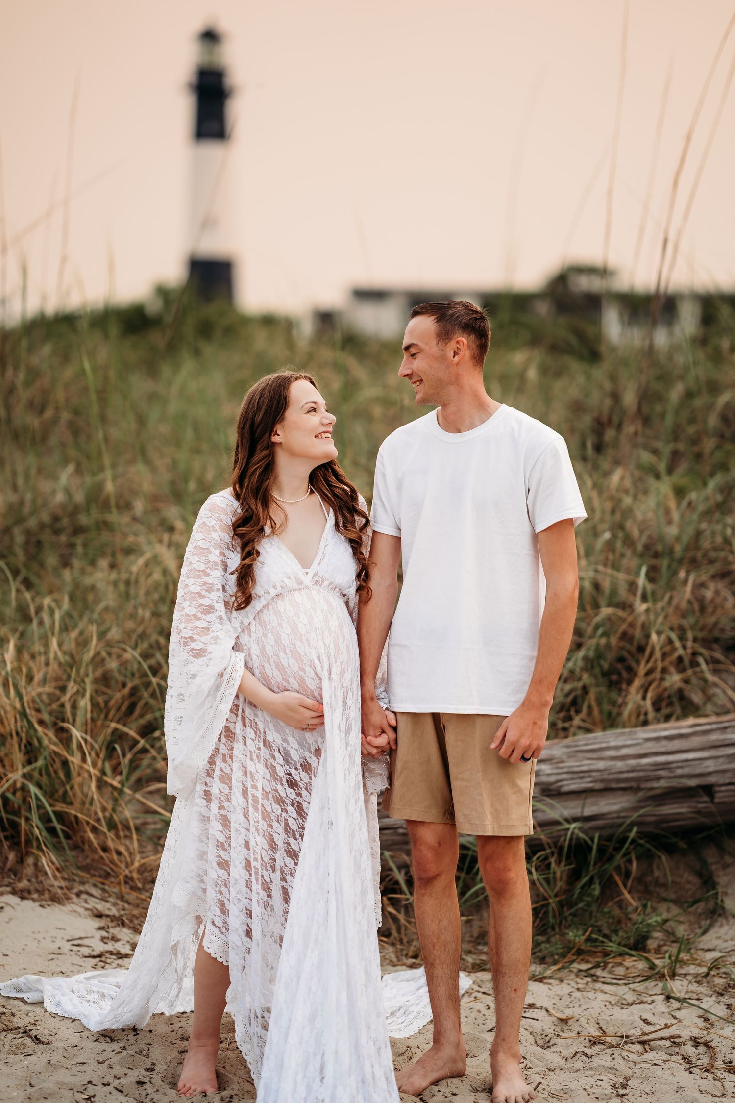 Smiling couple holding hands during maternity beach photoshoot with lighthouse visible in background.