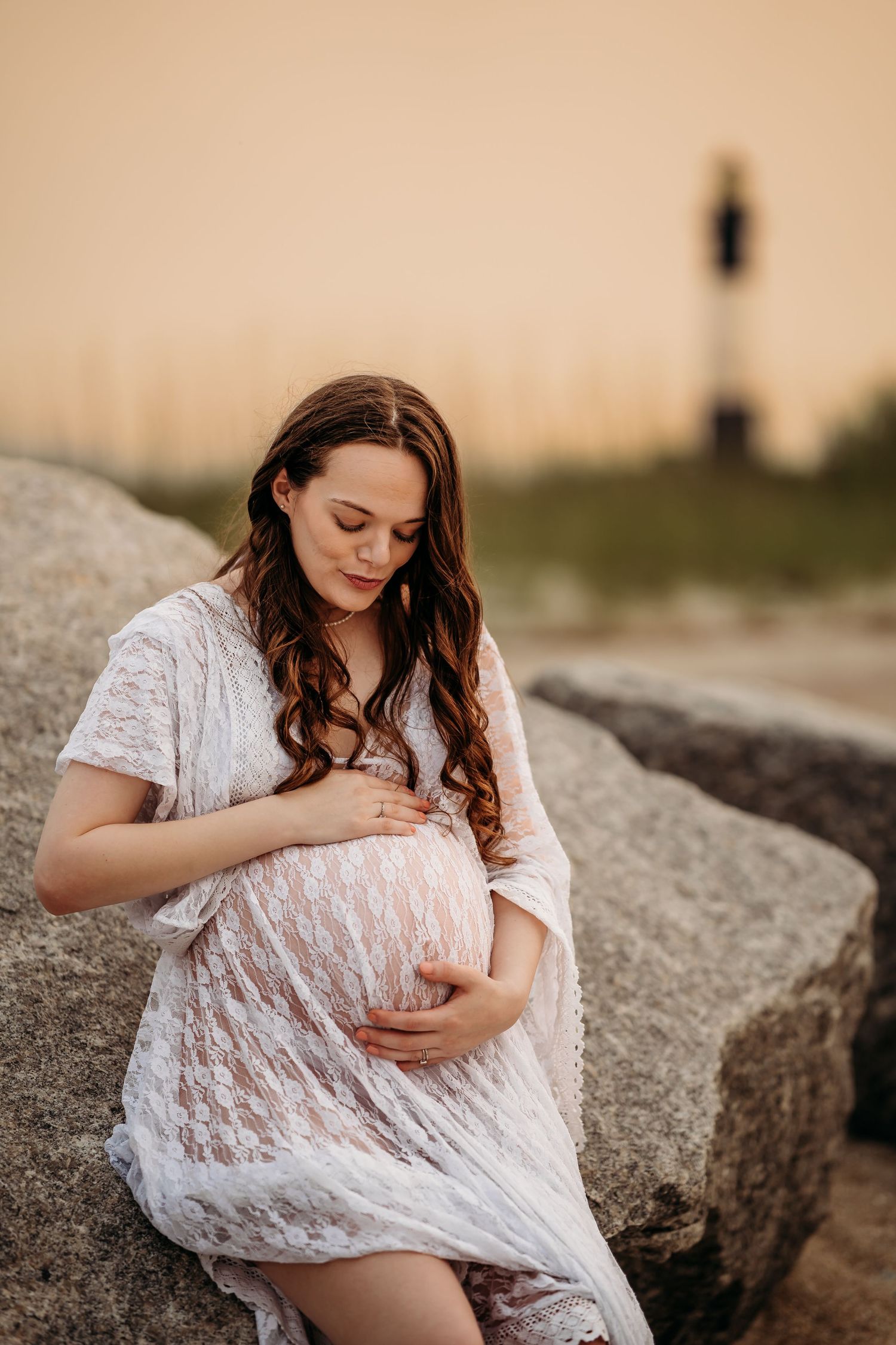 Pregnant woman in white lace dress sitting on rocks at sunset, gently cradling baby bump near lighthouse.