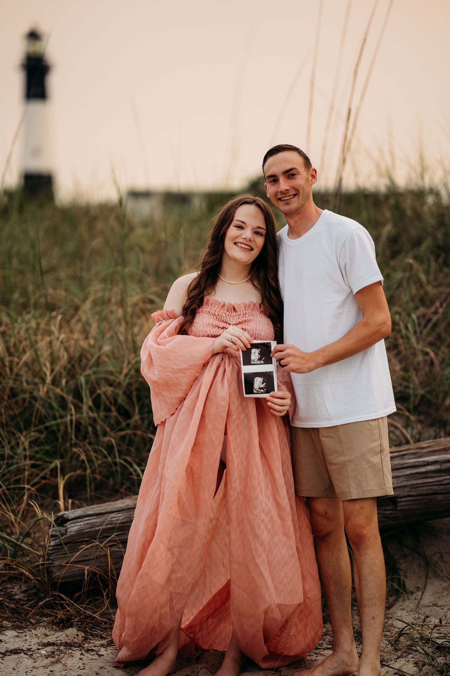 Smiling expecting couple holding ultrasound images near lighthouse and sea grass at sunset.