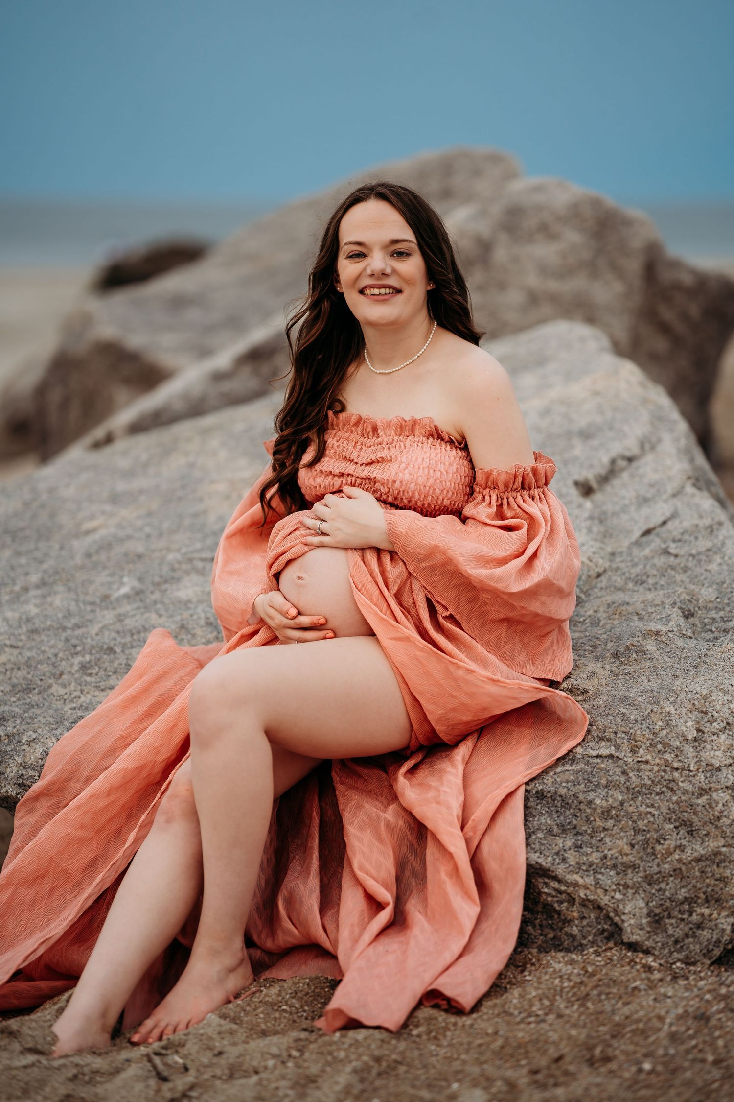Smiling pregnant woman in coral off-shoulder dress sitting on beach rocks, cradling her bump.