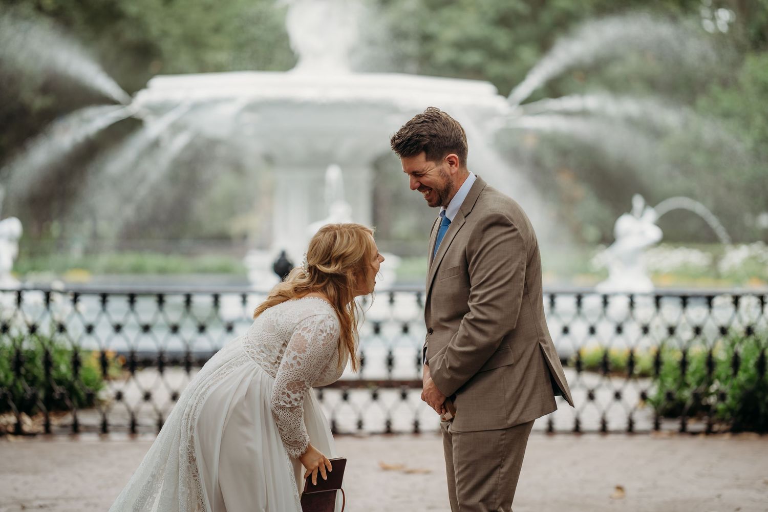 Bride in lace gown bows before groom in tan suit during outdoor wedding ceremony near ornate fountain.
