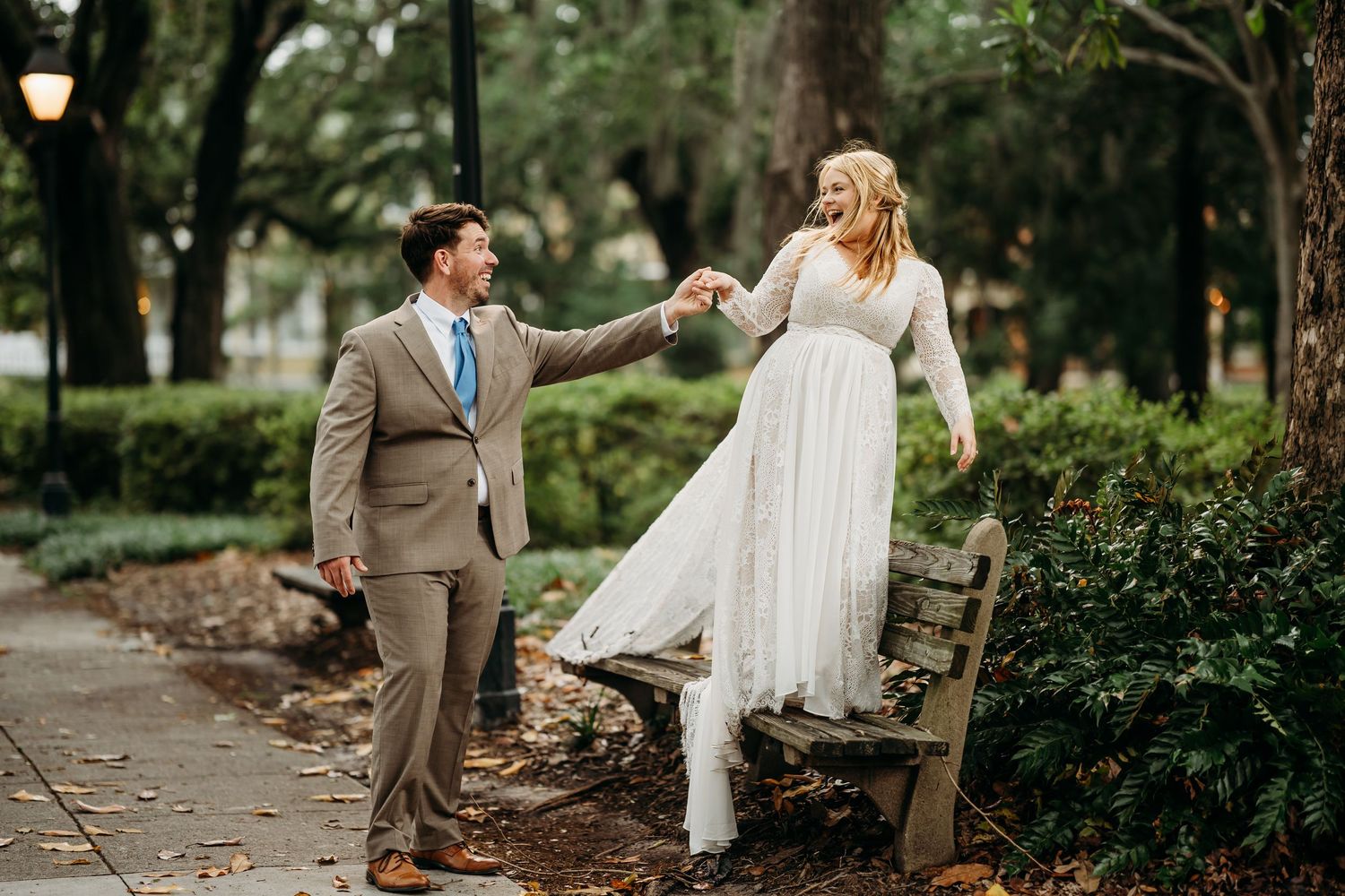 Bride and groom dance joyfully on a garden path, bride stepping off a bench in her flowing gown.