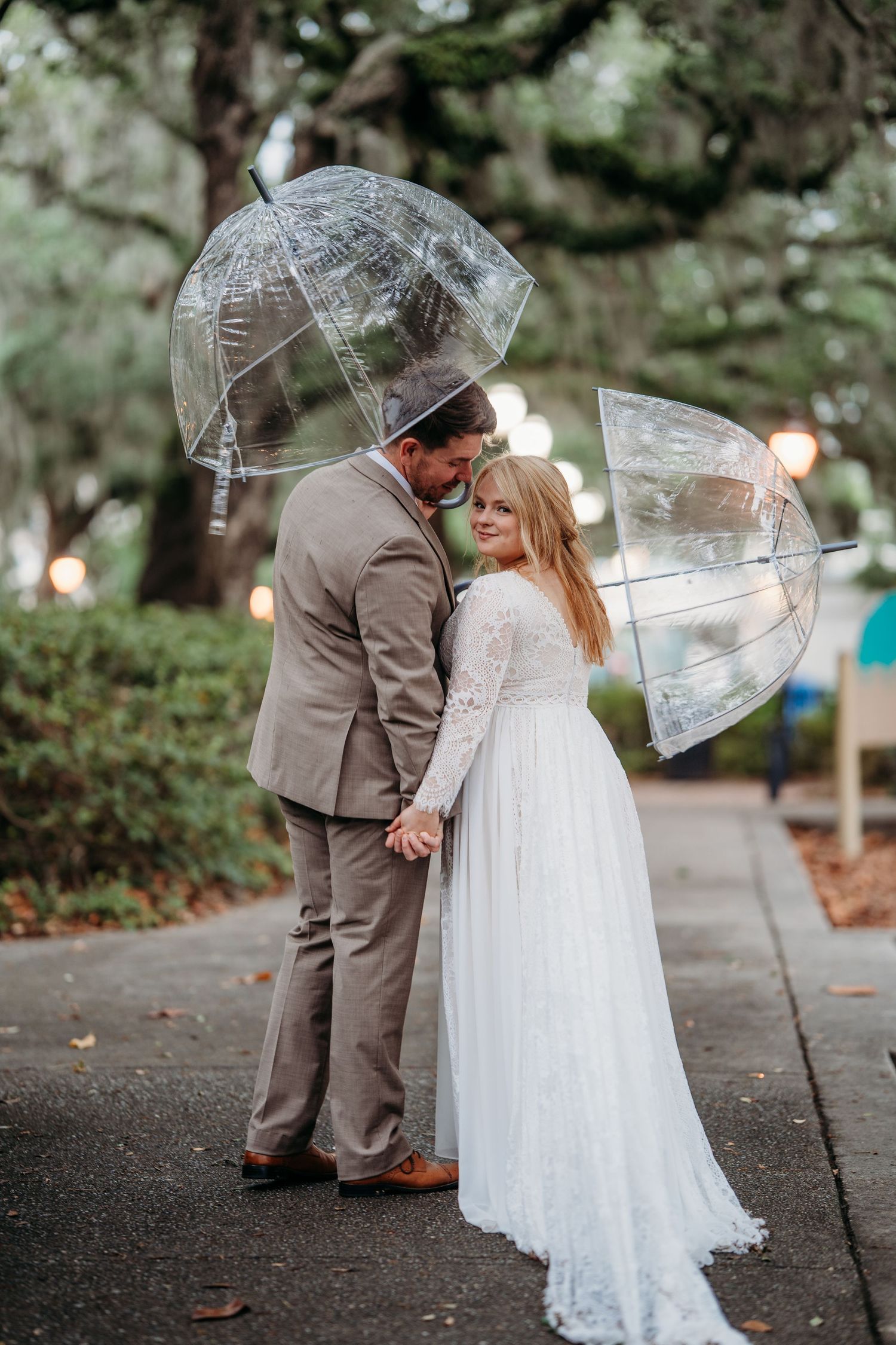 Bride in lace gown and groom in tan suit holding hands under clear umbrellas in a lush park.