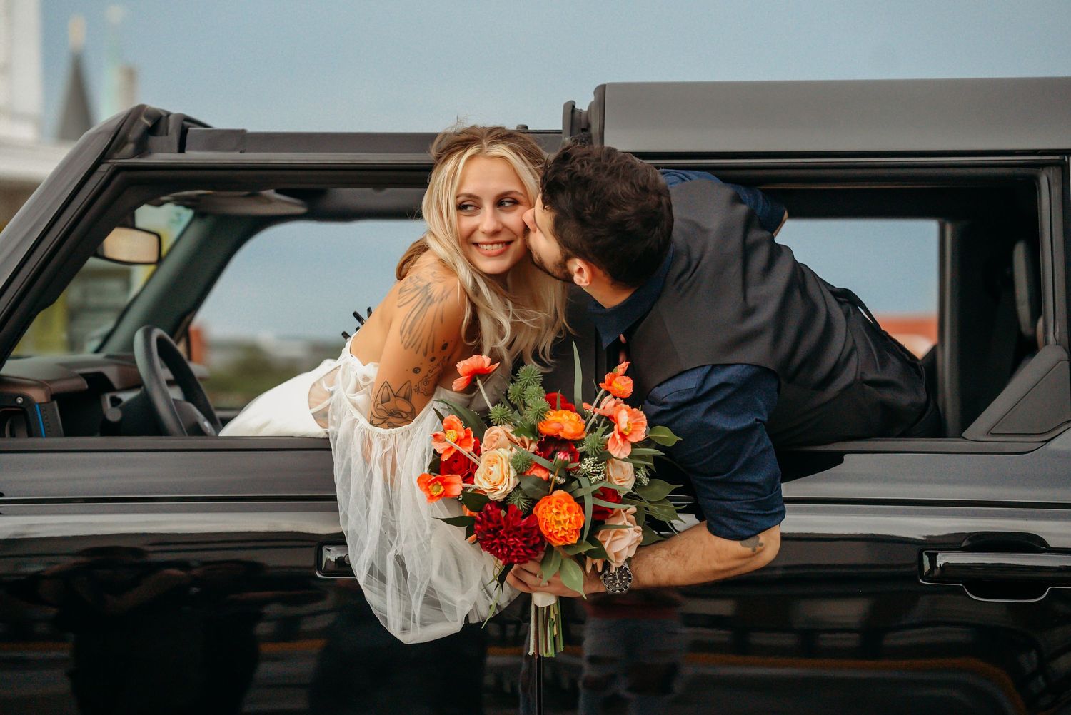 Woman smiling holding orange bouquet while man kisses her cheek by black truck.