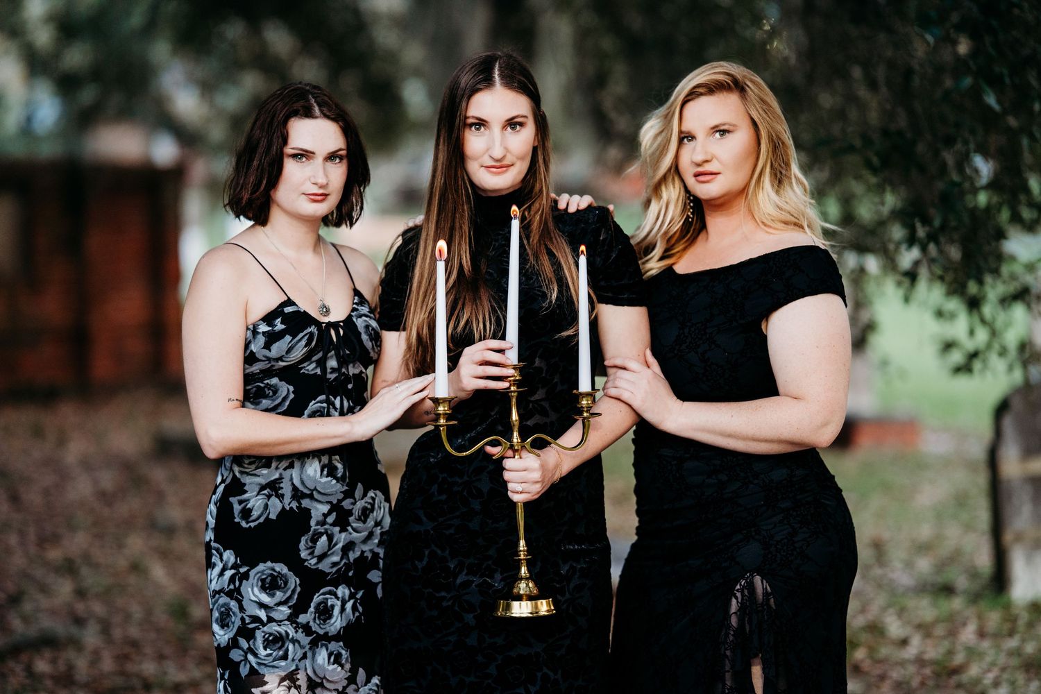 Three women in elegant black dresses posing outdoors with a candelabra in a gothic-style portrait.