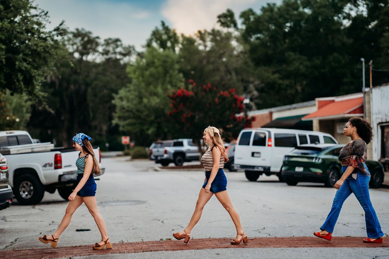 Three young women walking across a crosswalk in a small town, wearing summer outfits and sandals.