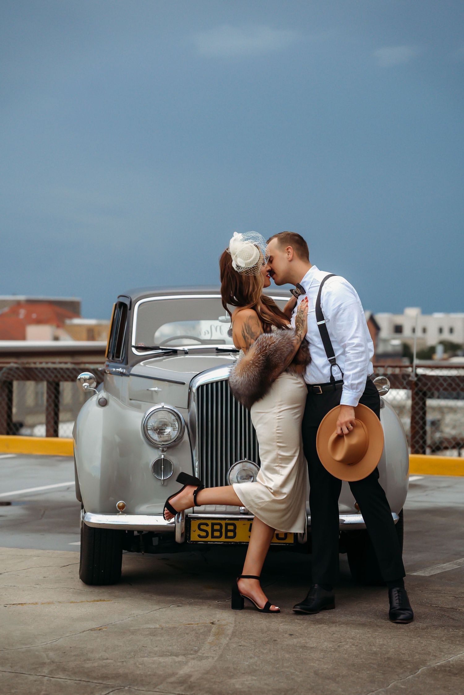 Couple kissing beside a vintage silver car on a rooftop parking lot, dressed in elegant retro wedding attire.