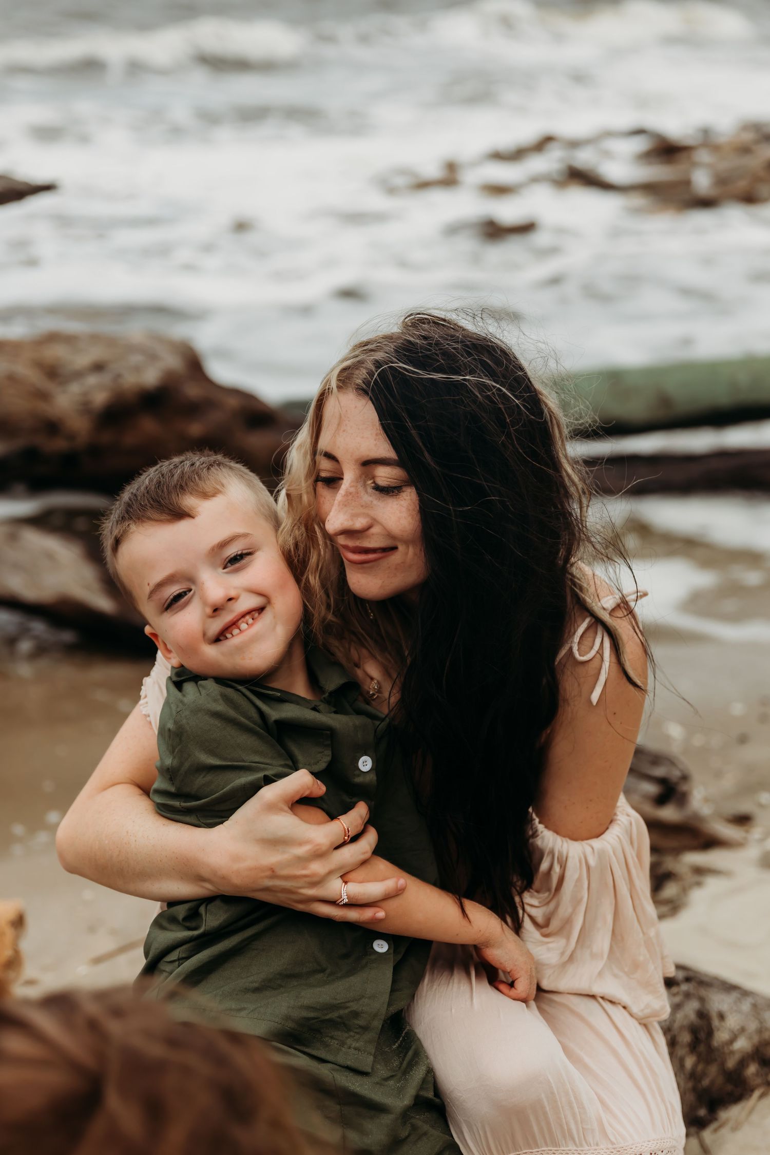 A woman with dark hair hugs a smiling young boy on a rocky beach, both dressed casually near ocean waves.