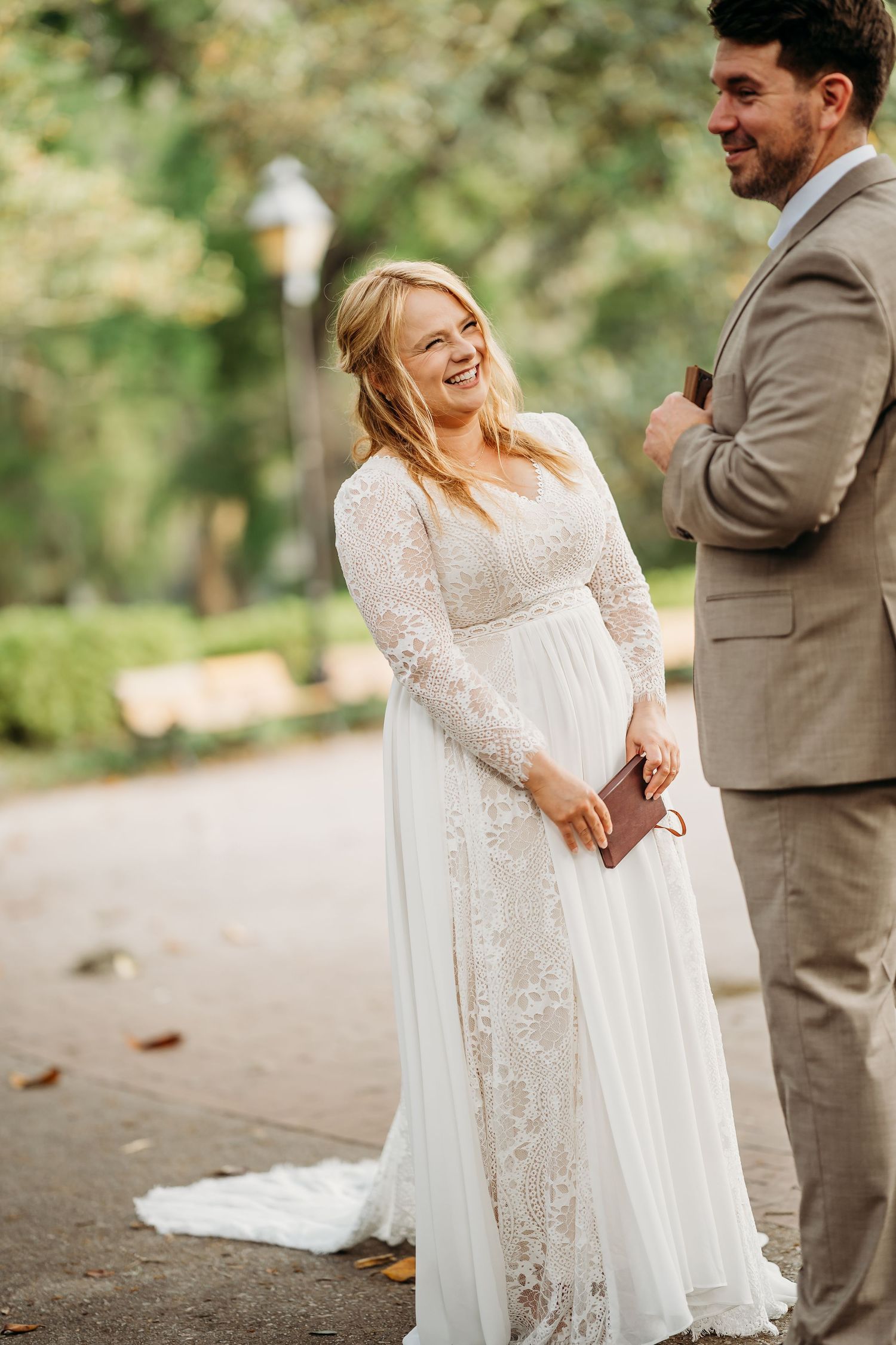 Smiling bride in lace long-sleeve wedding dress laughs joyfully with groom during outdoor ceremony.