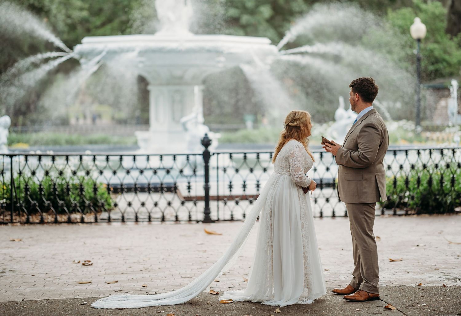 Bride and groom stand facing a grand fountain as groom reads vows during outdoor wedding ceremony.
