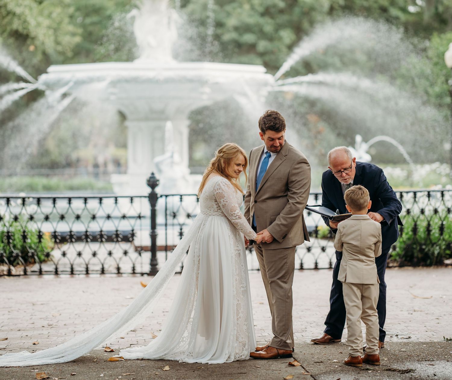 Bride and groom hold hands with officiant and child during intimate fountain wedding ceremony.