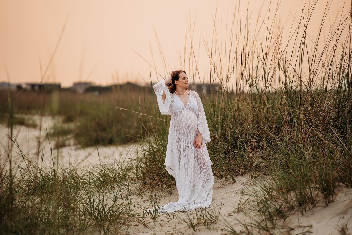 Pregnant woman in elegant white lace gown posing among tall sea grass on sandy beach at dusk.