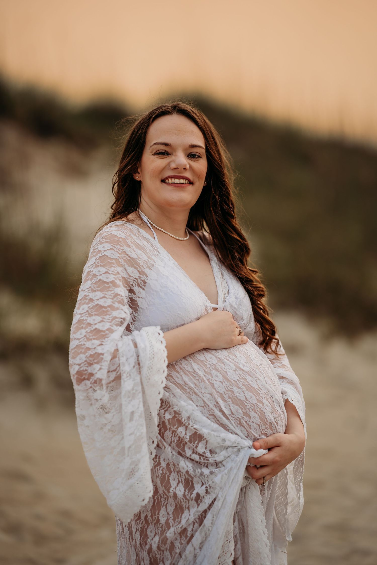 Smiling pregnant woman in white lace maternity gown standing on sandy beach at sunset.
